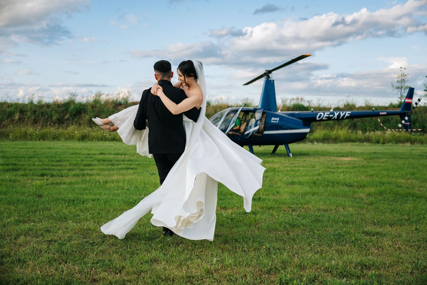 Groom lifting bride in white dress on green field with helicopter in background at outdoor wedding.