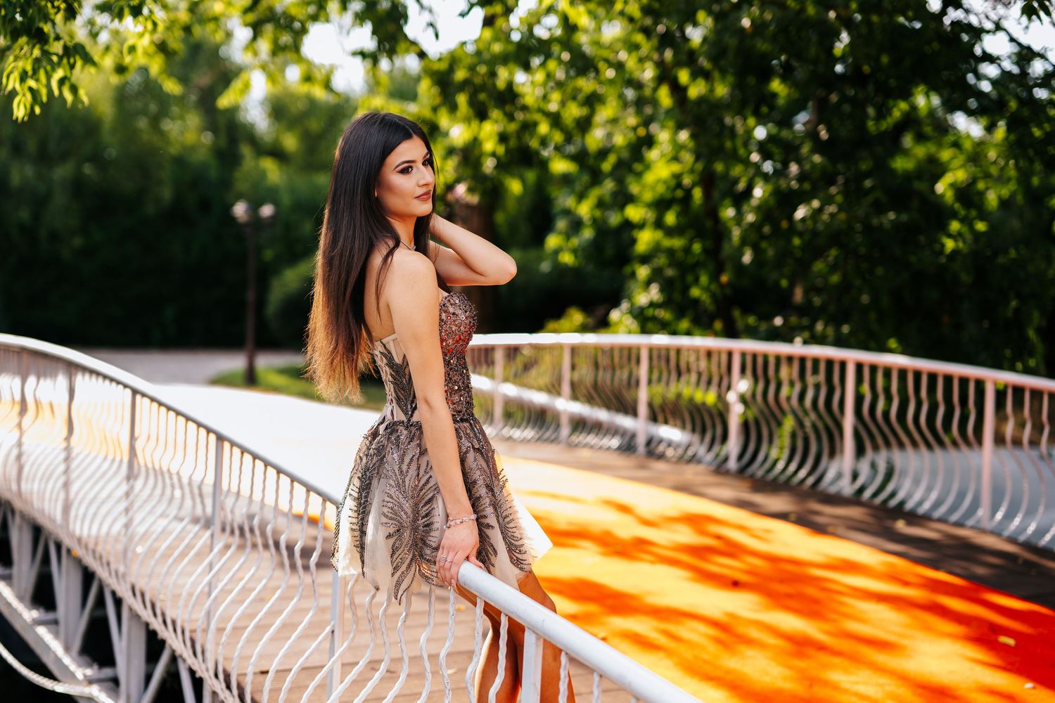 Young woman in elegant patterned dress posing on a white bridge railing in a lush green park at sunset.