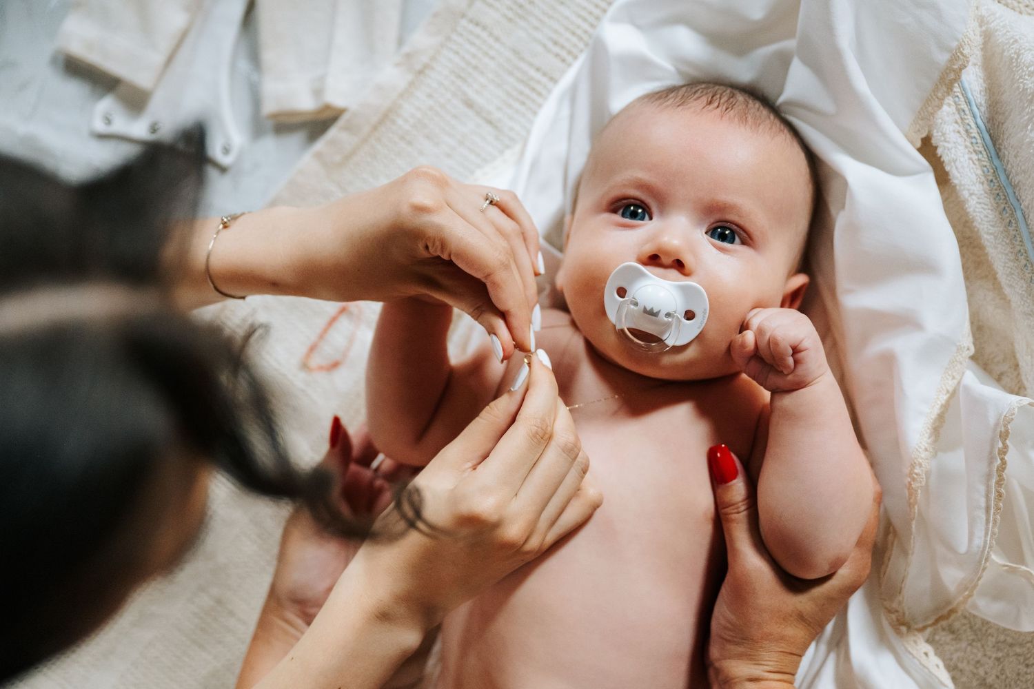A mother with red nails changes her newborn baby's diaper, as the infant looks up with a pacifier.