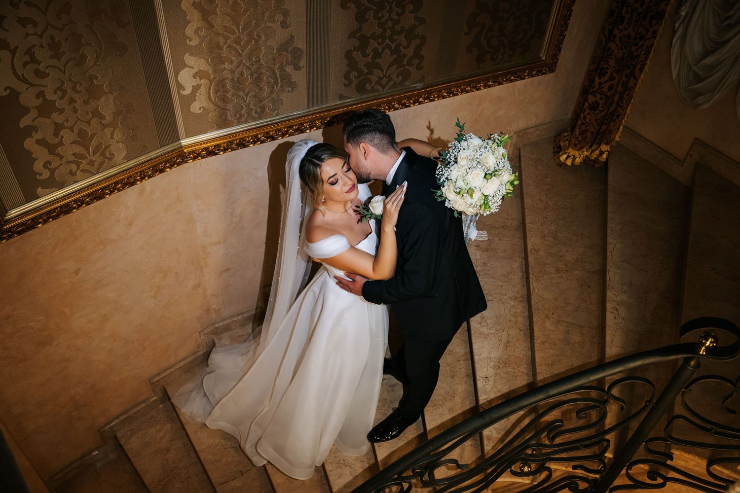 Bride and groom embrace on a grand ornate staircase, bride holding a white bouquet, viewed from above.