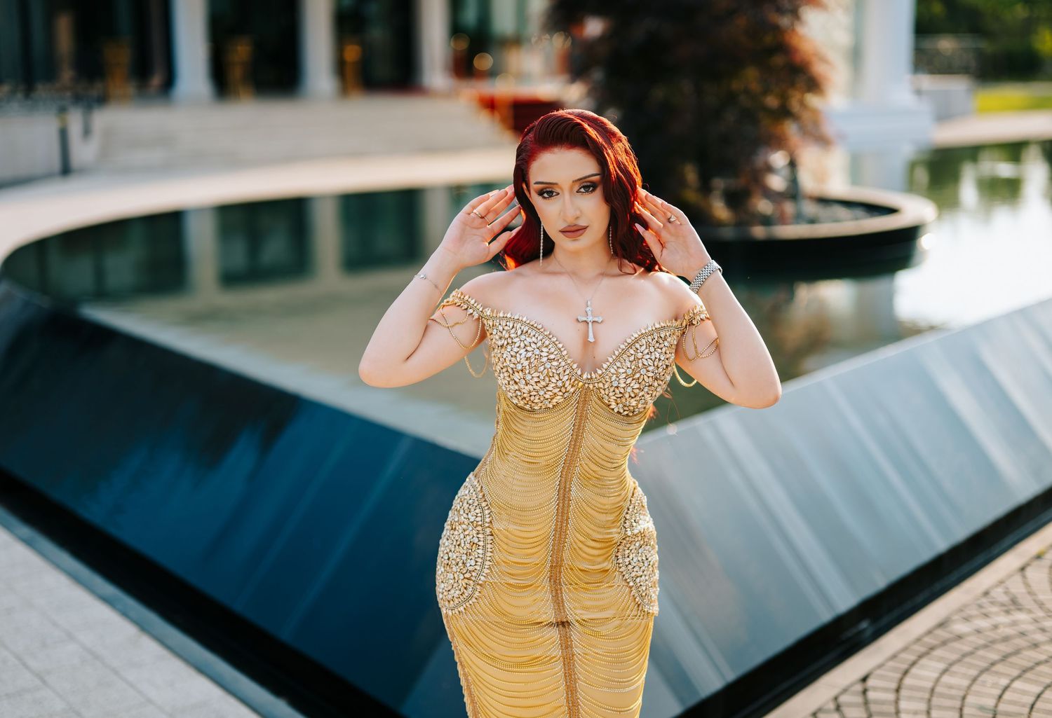 Woman in gold embellished form-fitting gown posing near a fountain outdoors.