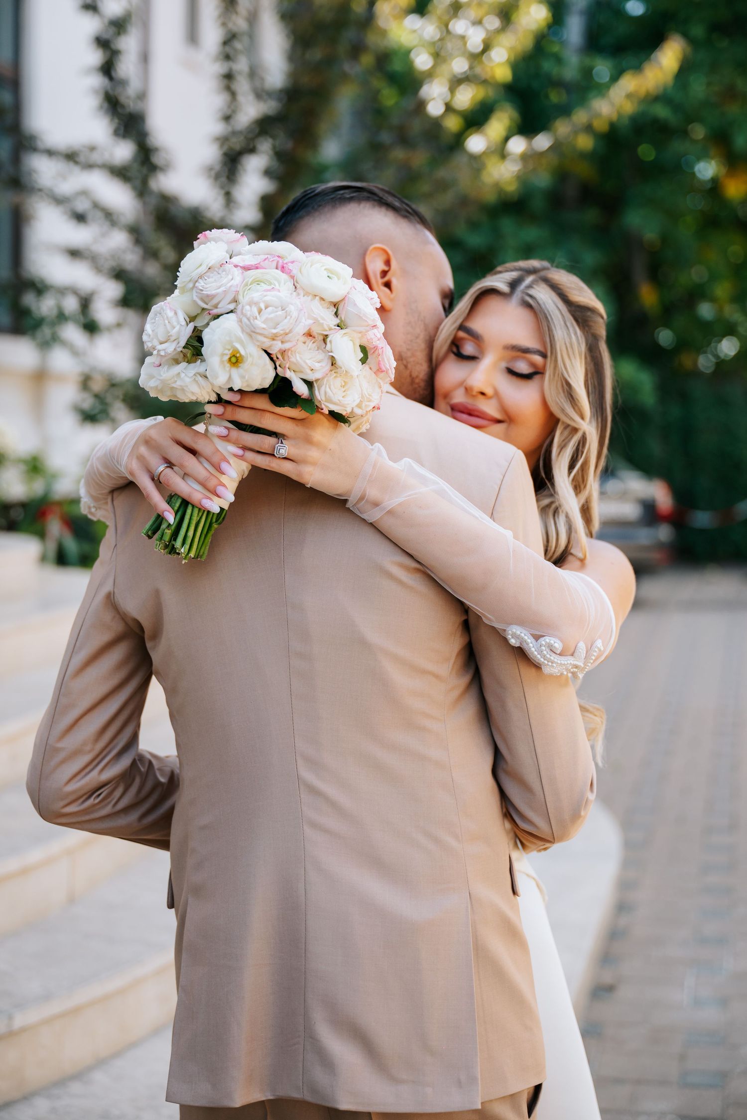 Bride in white dress hugging groom in beige suit while holding white peony bouquet outdoors.