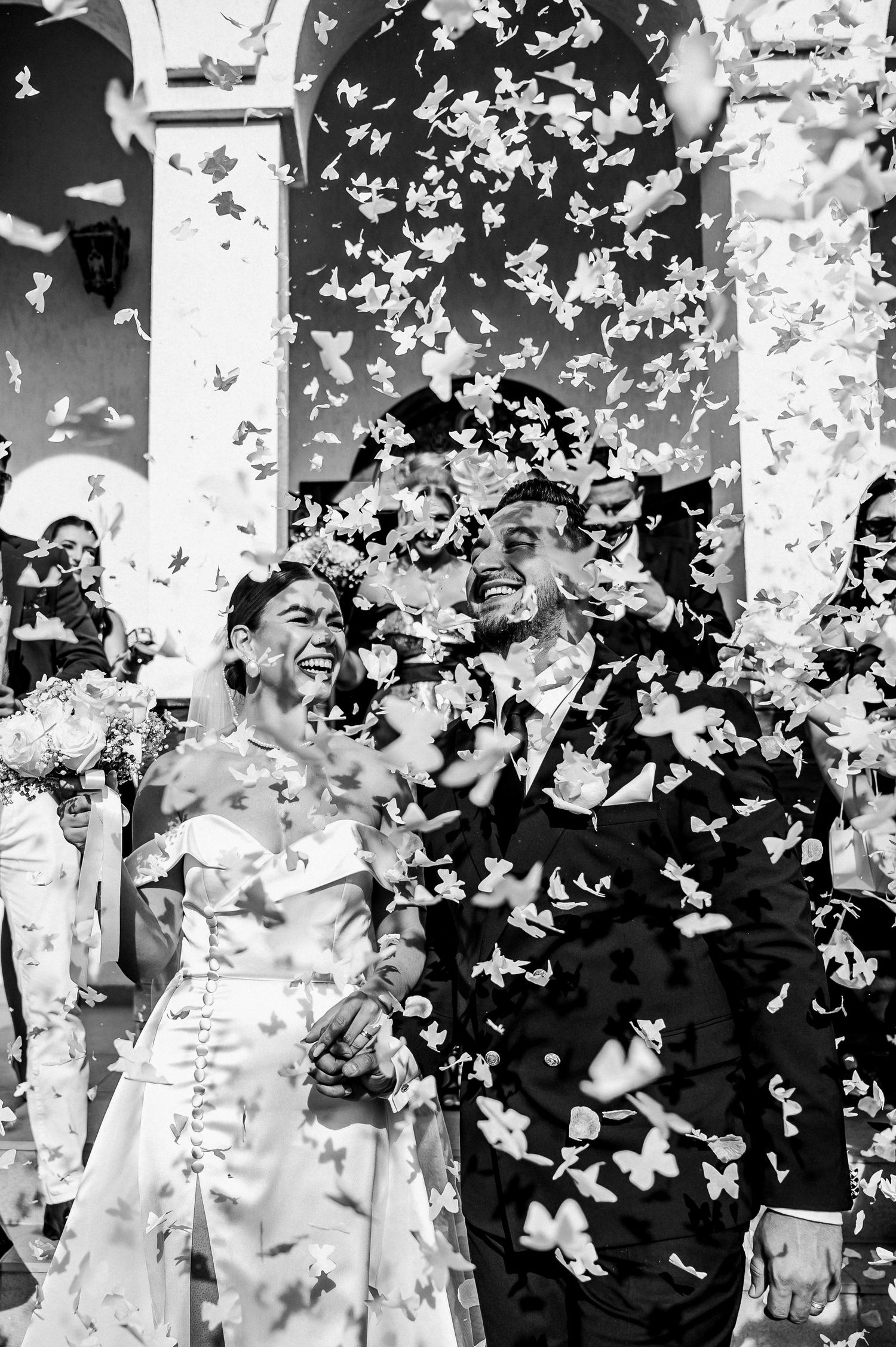 Black and white photo of a joyful bride and groom laughing as confetti fills the air outside a church.
