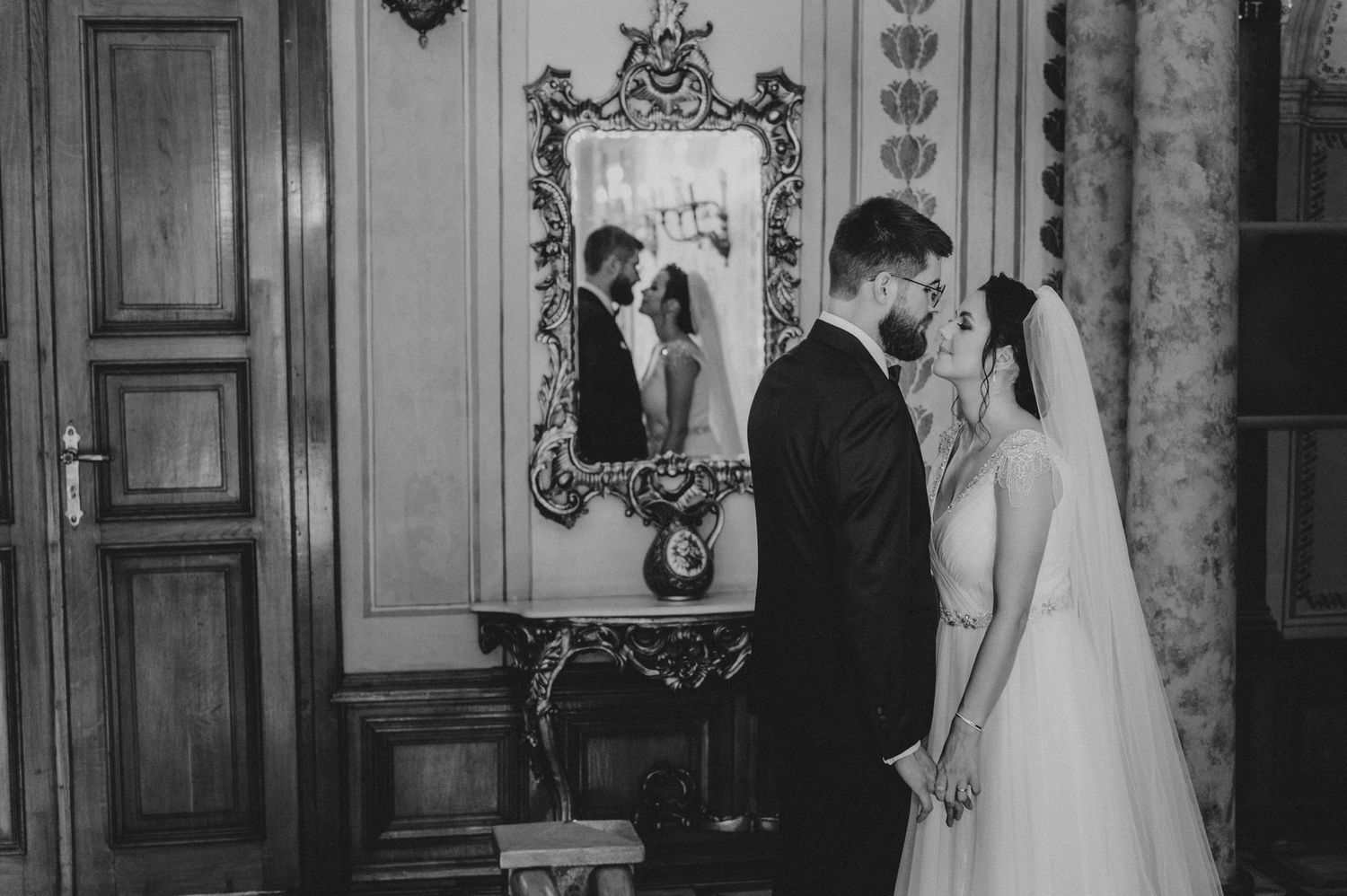 Black and white wedding photo of bride and groom sharing intimate moment, reflected in ornate vintage mirror.