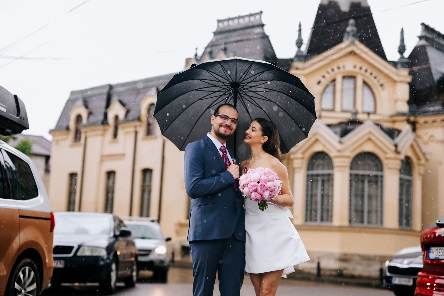 Newlyweds share a romantic moment under a black umbrella in the rain, bride holding pink bouquet in white dress.