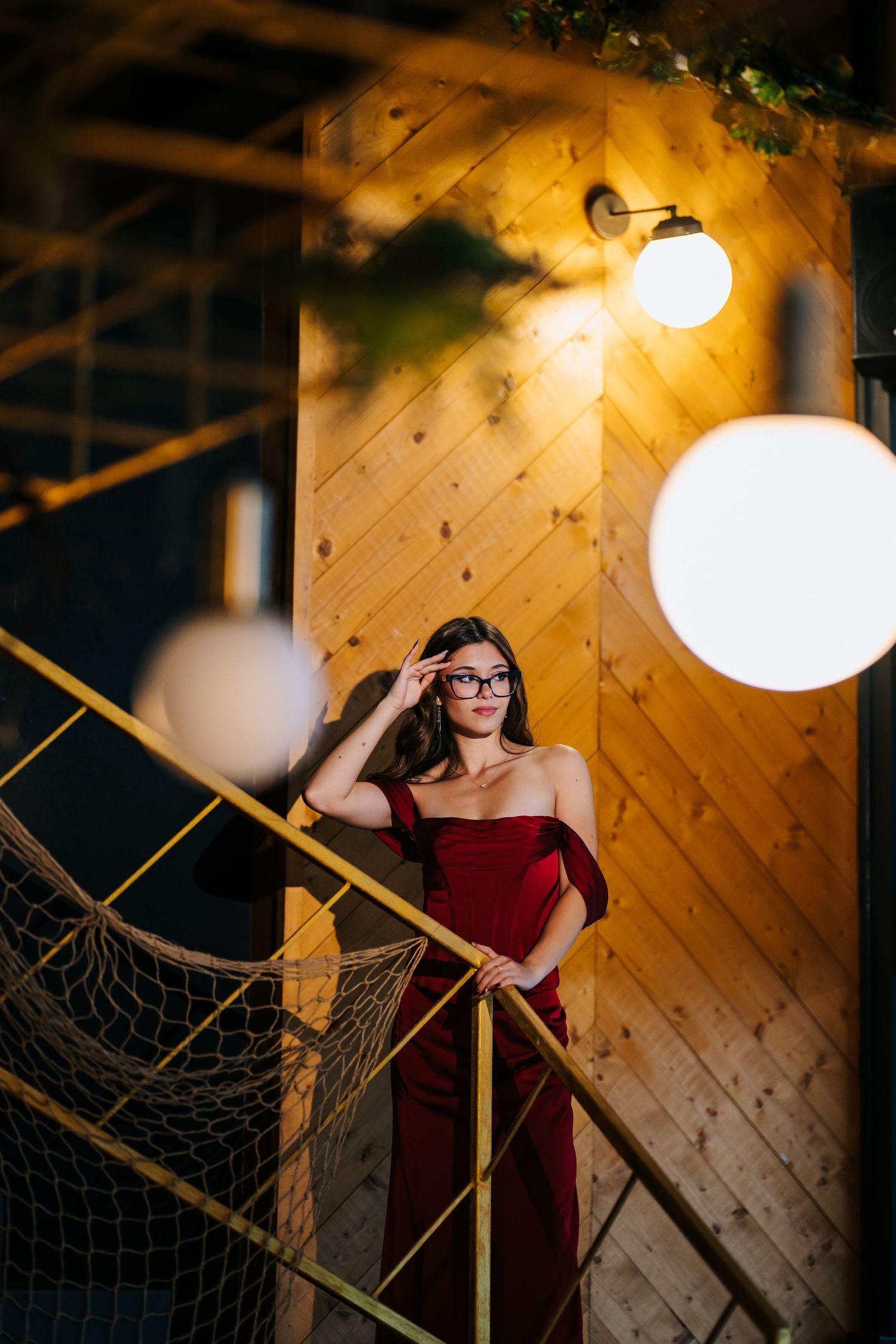 Woman in red off-shoulder dress and glasses poses on staircase with warm glowing lights and wooden wall backdrop.