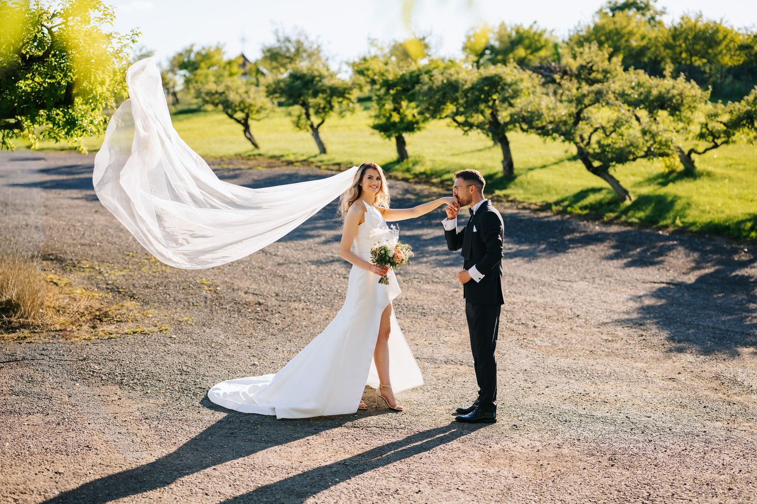 Bride in white gown with flowing veil and groom in black tuxedo pose on a sunlit path surrounded by lush green trees.