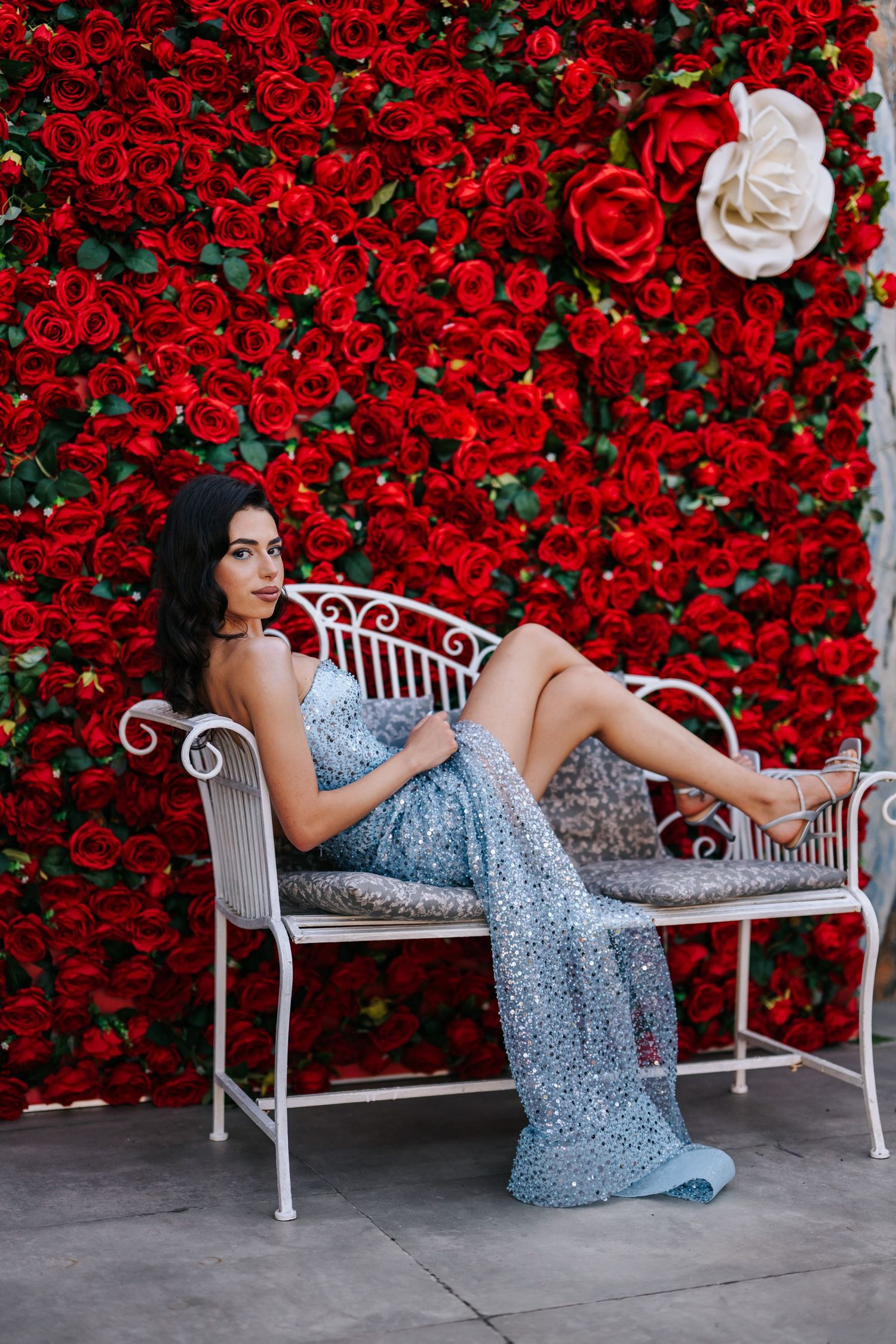 Woman in silver sequin gown poses on white bench against red rose wall backdrop.