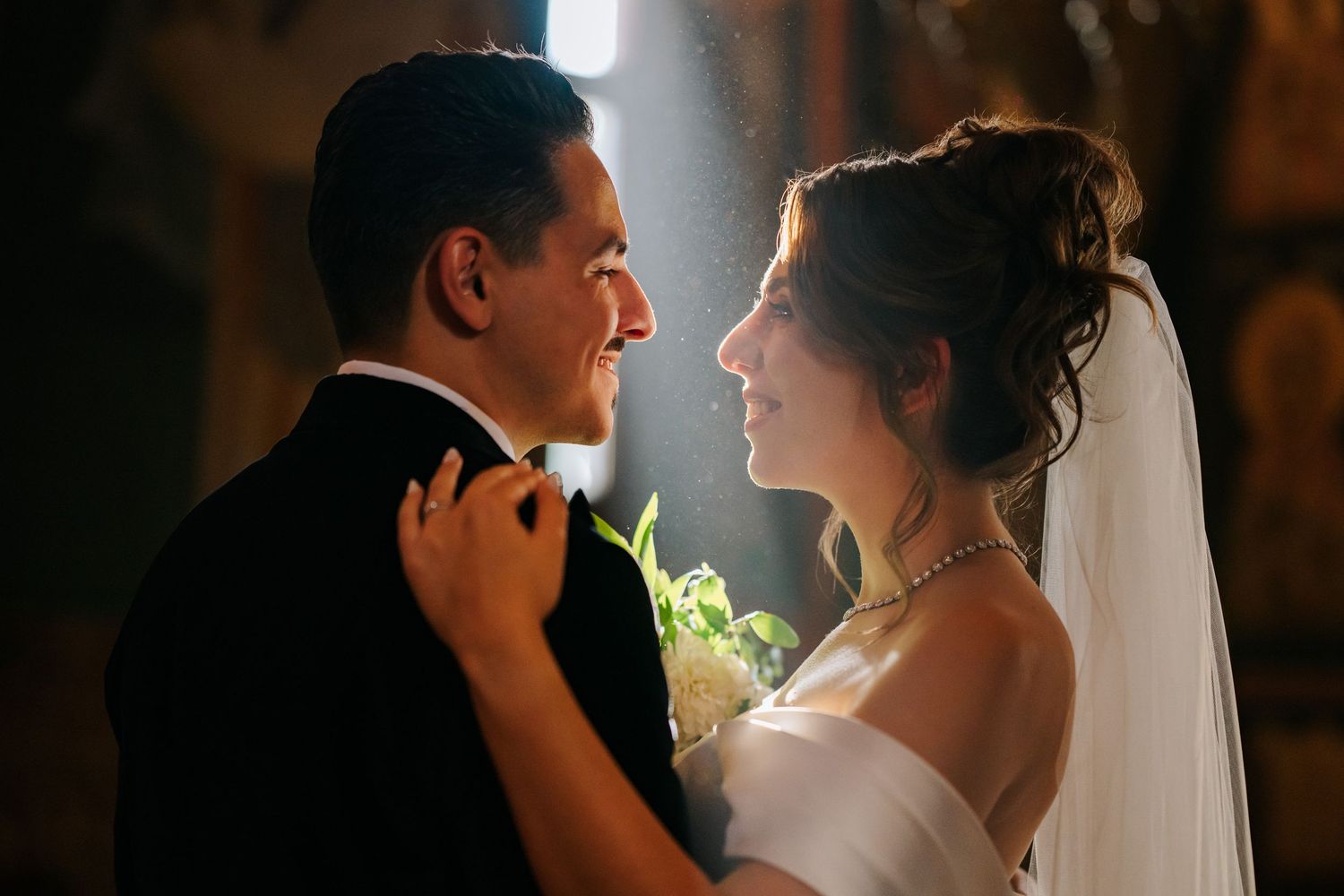 Bride and groom share a tender smile in a softly lit church, with the bride holding white flowers.