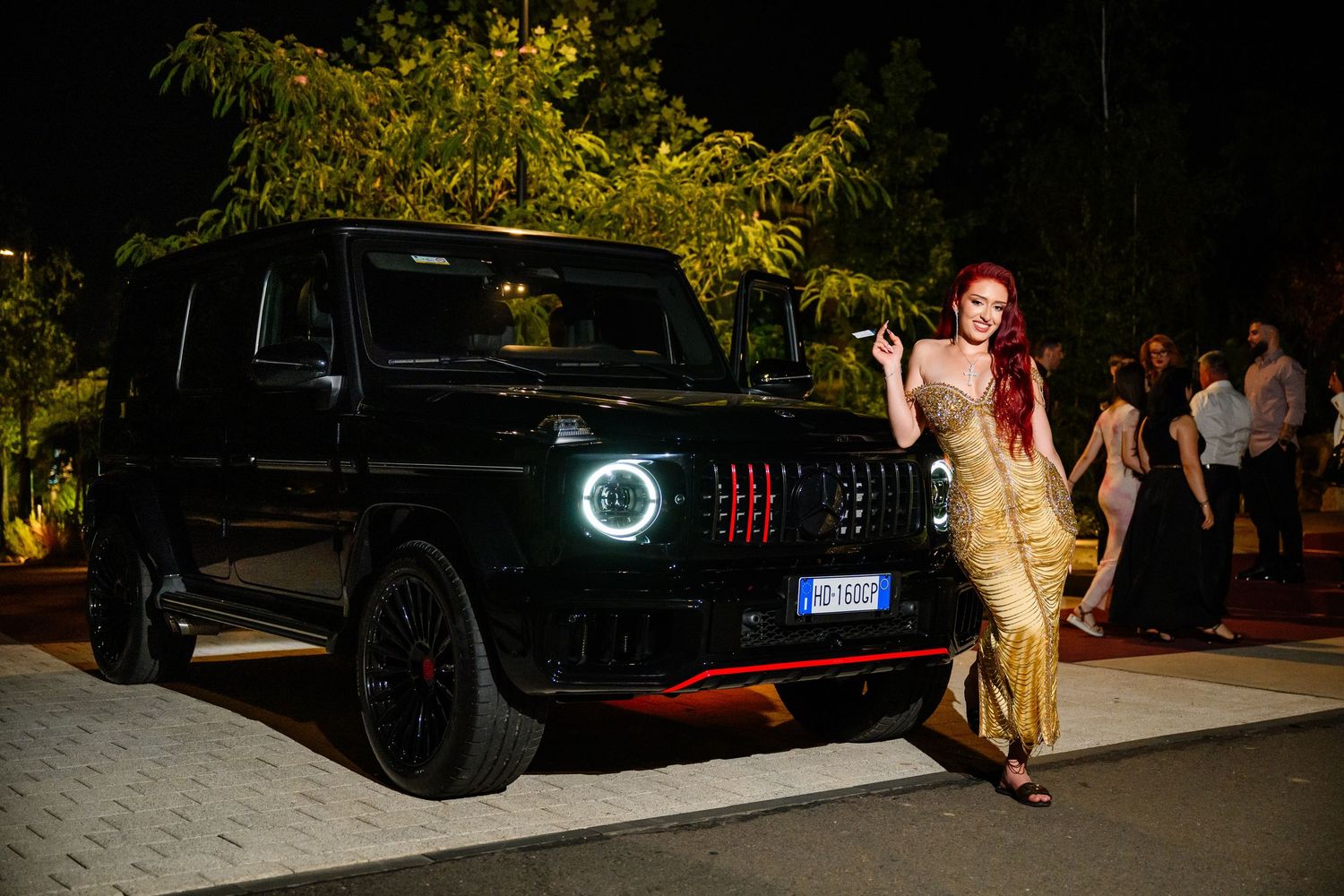Woman in gold dress posing beside black Mercedes-AMG G-Wagon at night event.