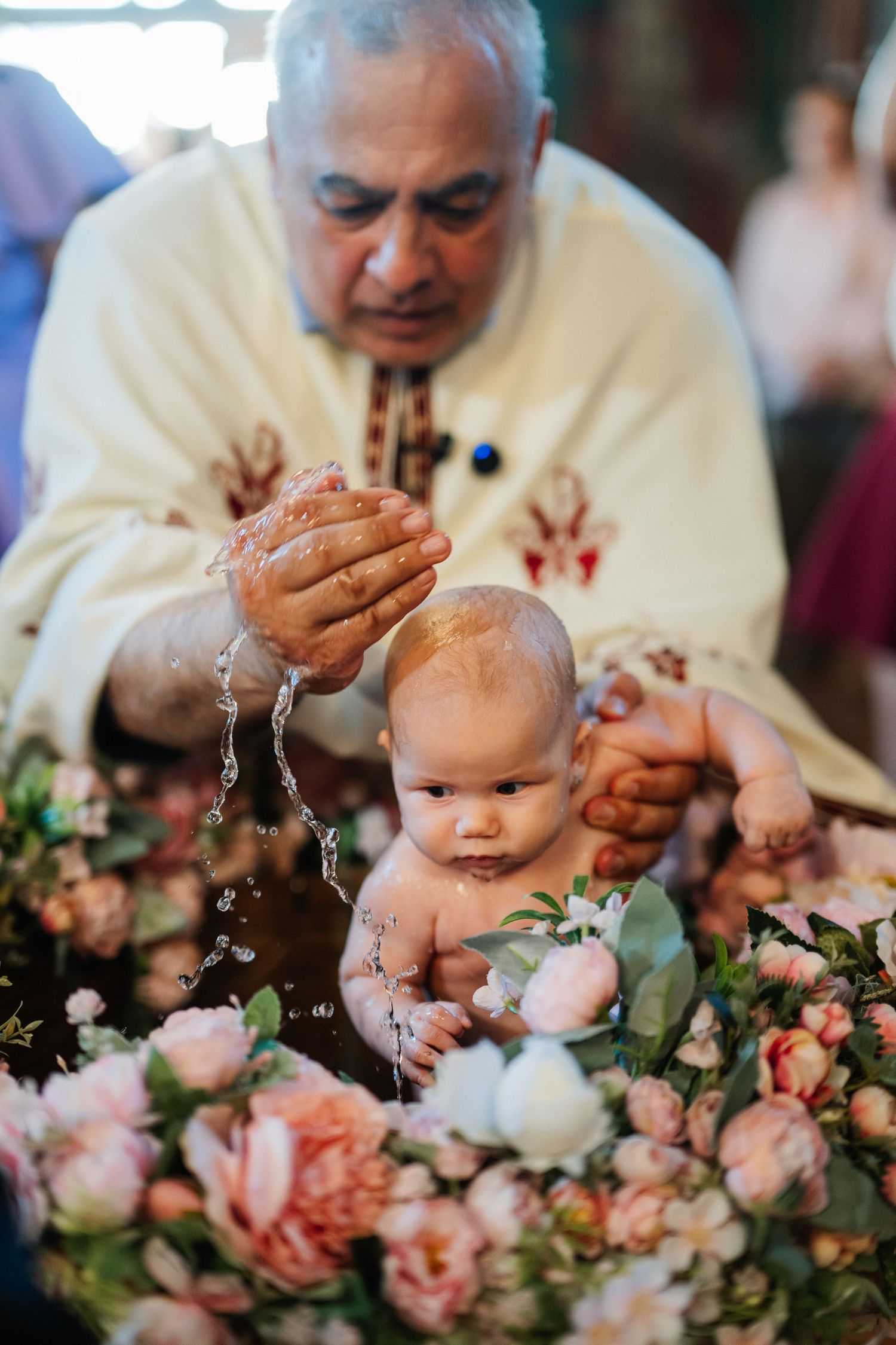 Orthodox priest pours holy water over a baby during a traditional baptism surrounded by pink flowers.