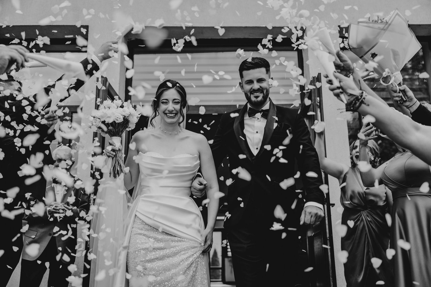 Black and white photo of a joyful newlywed couple walking through a shower of confetti at their wedding ceremony.