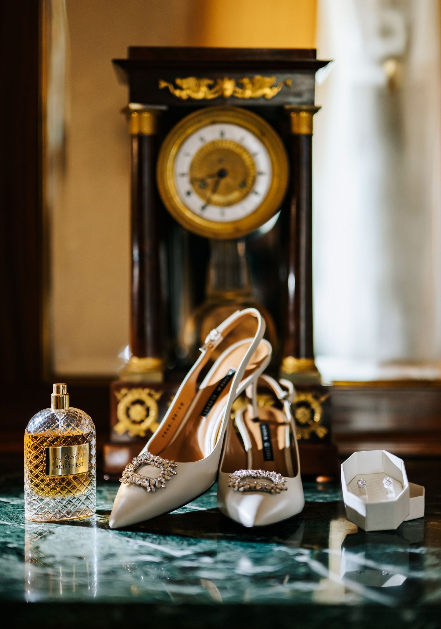 Elegant bridal heels with crystal buckles displayed on marble beside perfume and an antique clock.