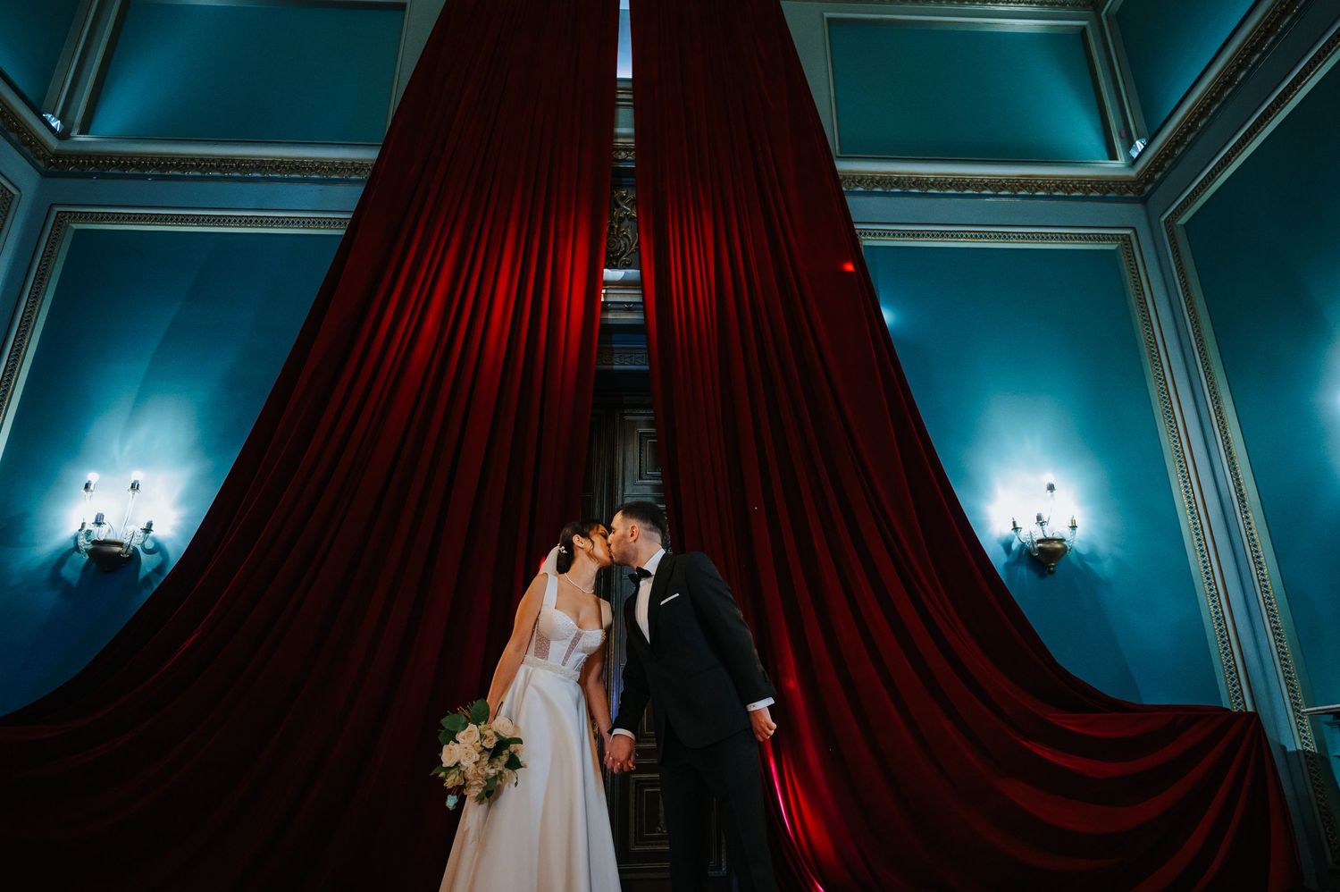 Bride and groom kissing in front of dramatic red velvet curtains in an elegant blue-walled wedding venue.