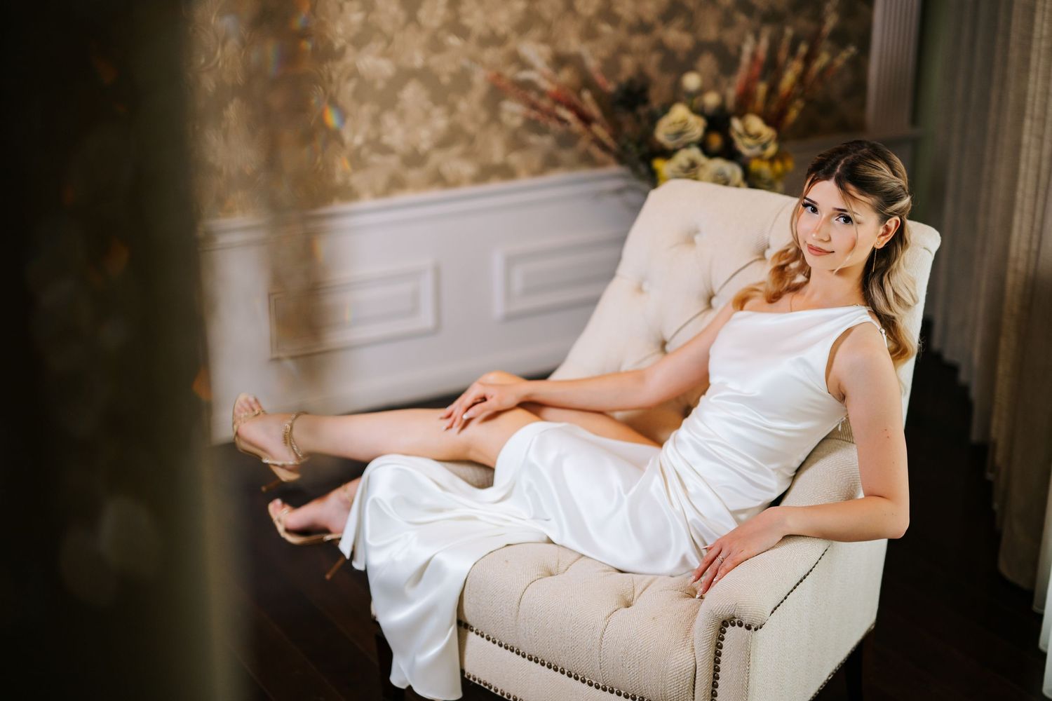 Elegant bride in white gown relaxing in cream tufted chair, wearing floral headpiece in ornate room.