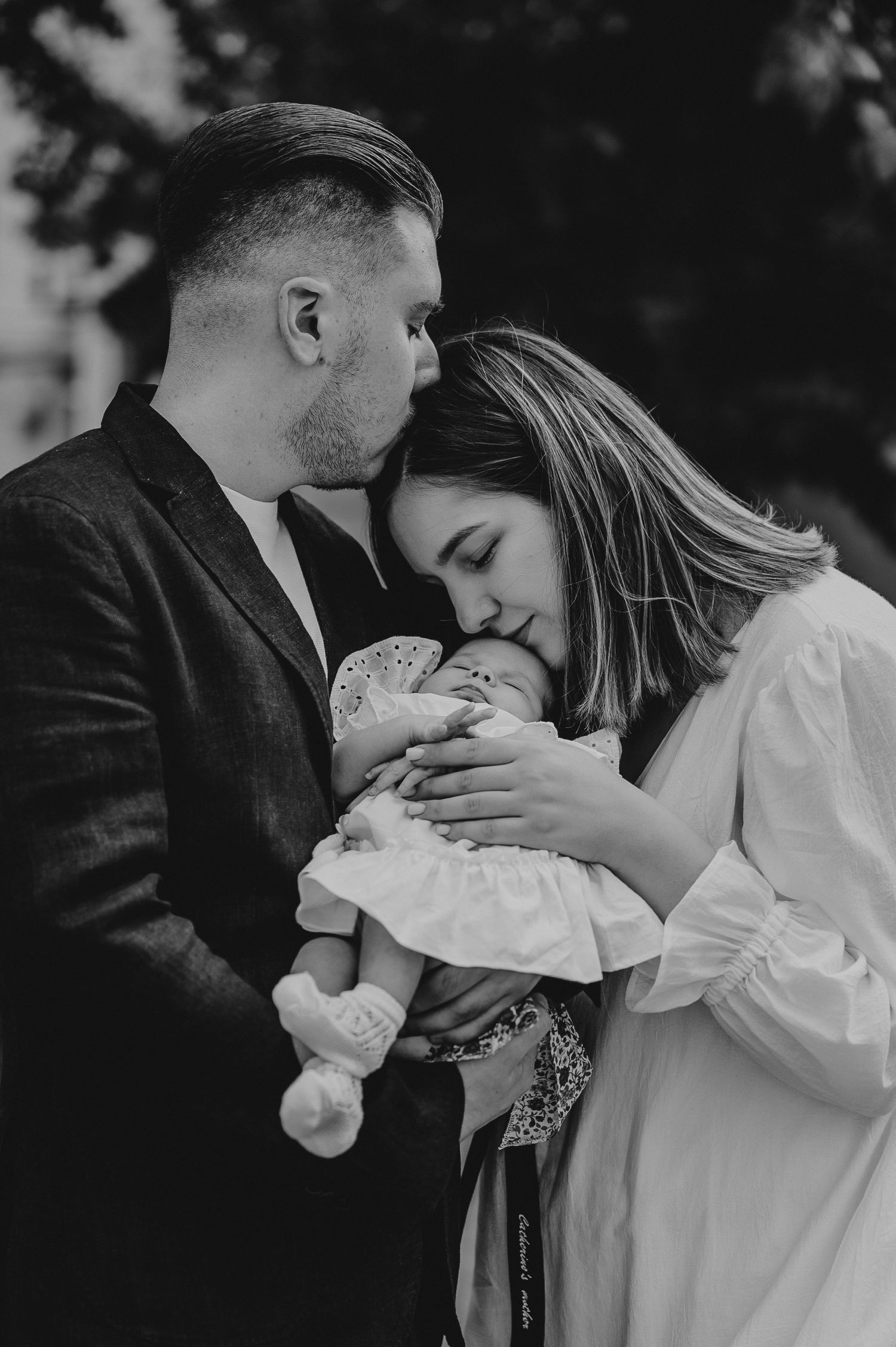 Black and white photo of a couple tenderly holding their newborn baby outdoors, sharing an intimate family moment.