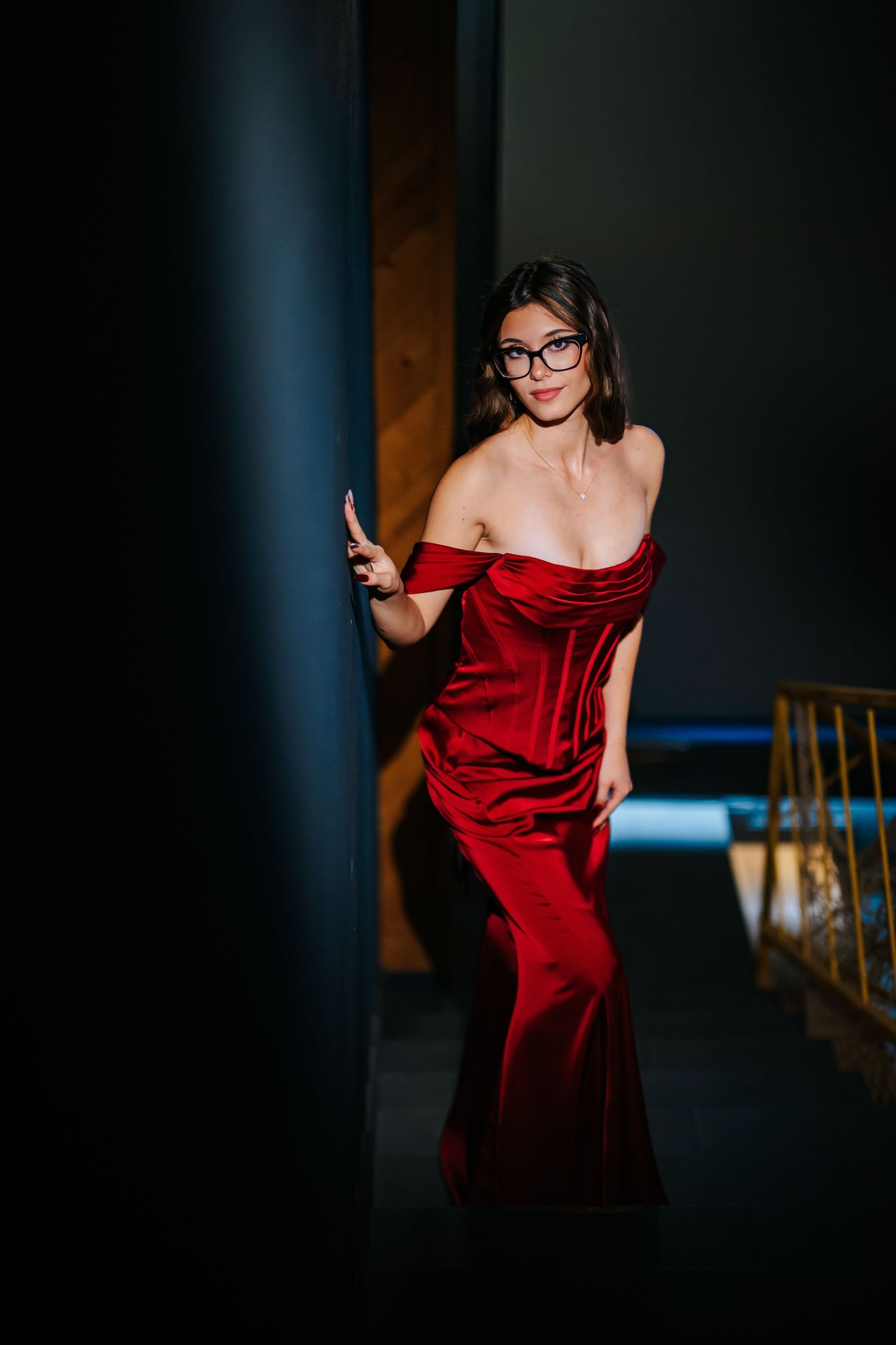 Elegant woman in red off-shoulder velvet gown and glasses posing in dramatic lighting.
