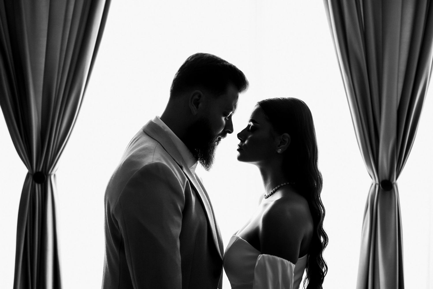 Black and white silhouette of a romantic couple facing each other framed by elegant curtains in a wedding portrait.