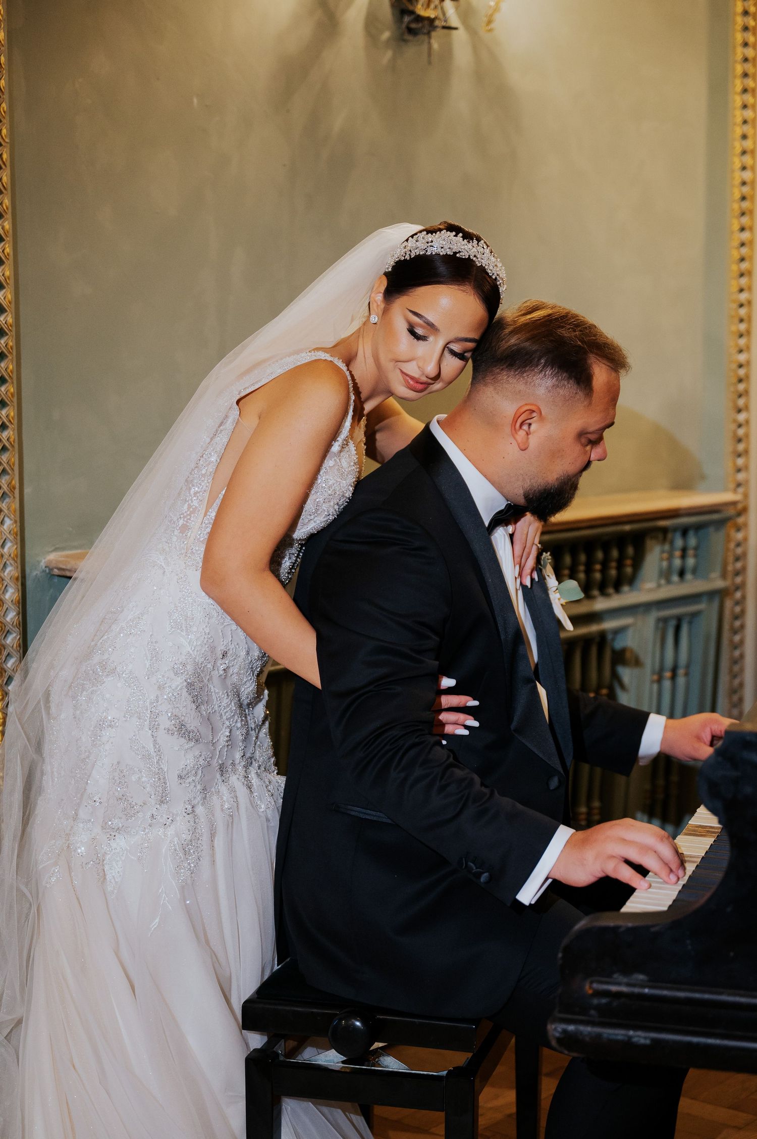Bride in white gown and tiara leans over groom in tuxedo playing piano at elegant wedding venue.