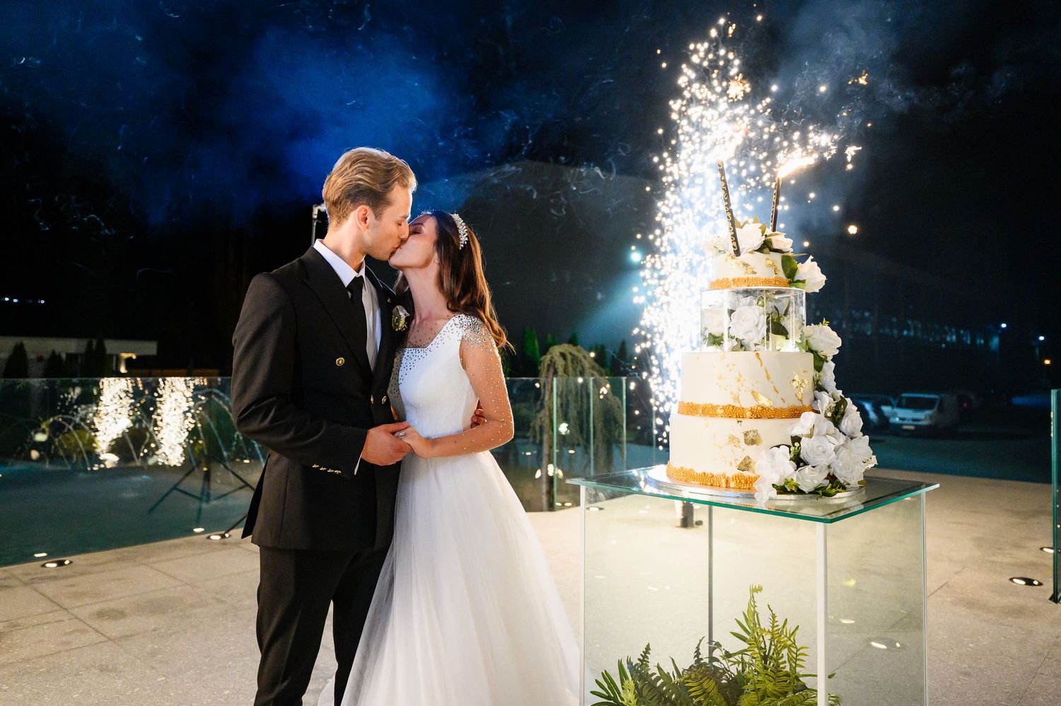 Bride and groom share a kiss at night with sparkling fireworks and white wedding cake backdrop.