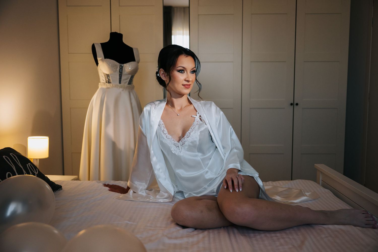Woman in white lingerie sitting on bed with elegant wedding dress on mannequin in softly lit bedroom.