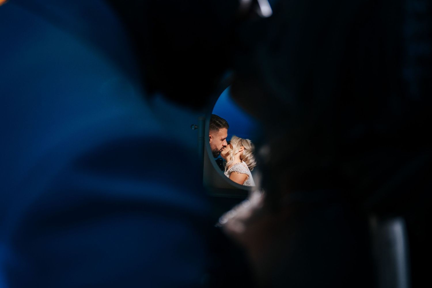 Couple shares an intimate kiss captured through a creative framed perspective in soft warm light.