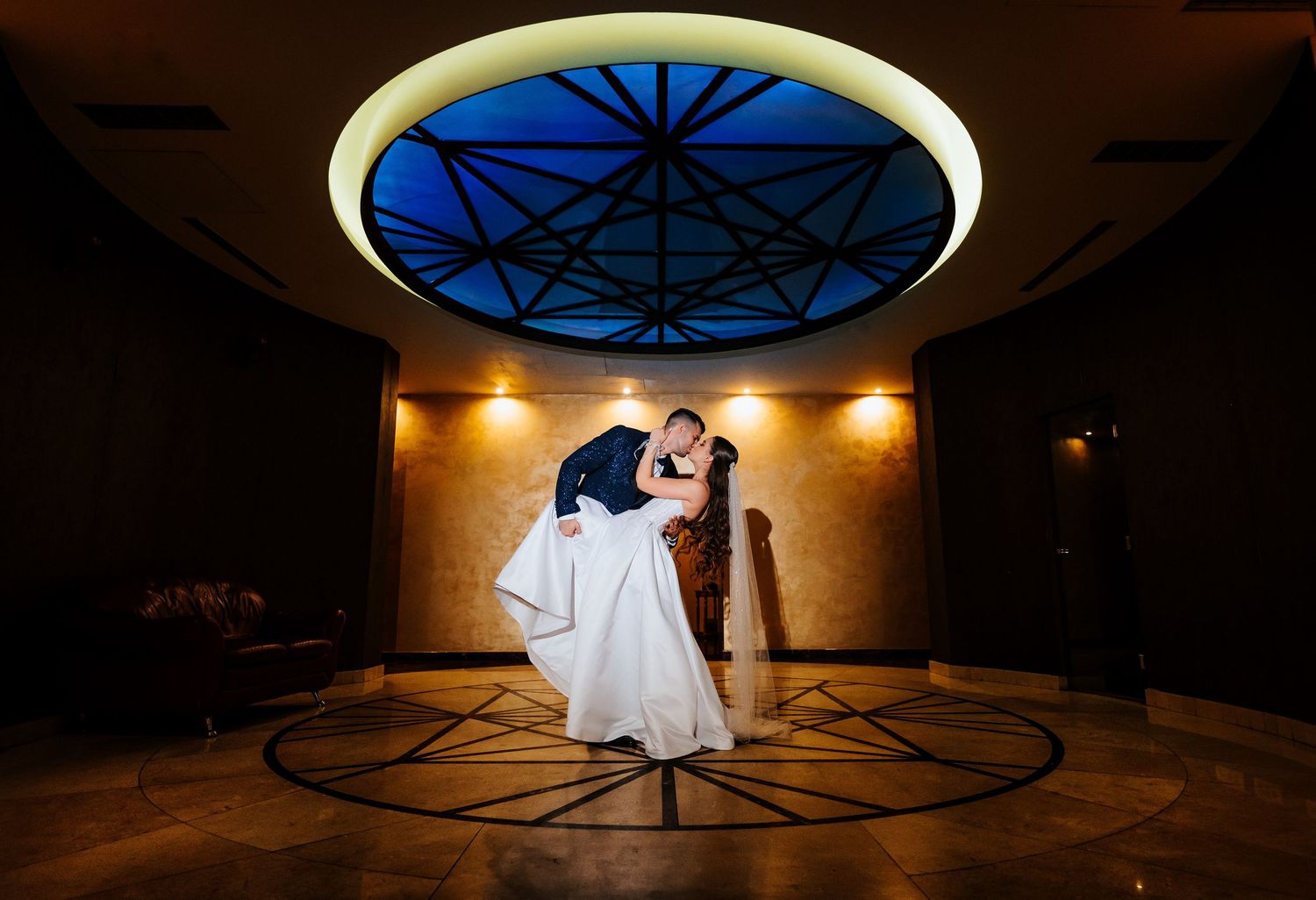 Groom dips bride under a stunning blue geometric ceiling light in an elegant wedding venue lobby.