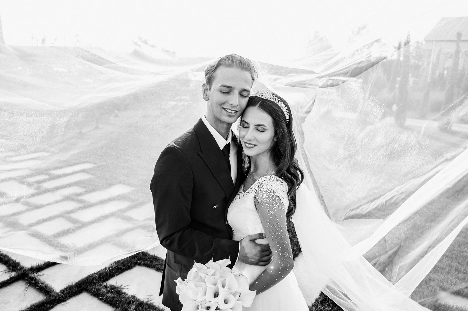 Black and white wedding photo of smiling bride and groom embracing outdoors, brides veil flowing in the wind.