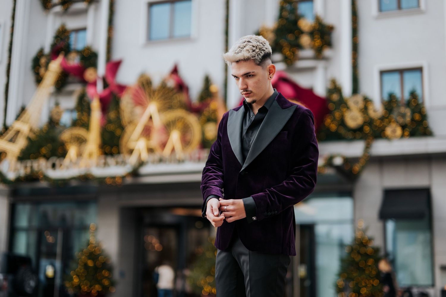 Stylish man in purple velvet blazer adjusting cufflinks outdoors near festive Christmas decorations.