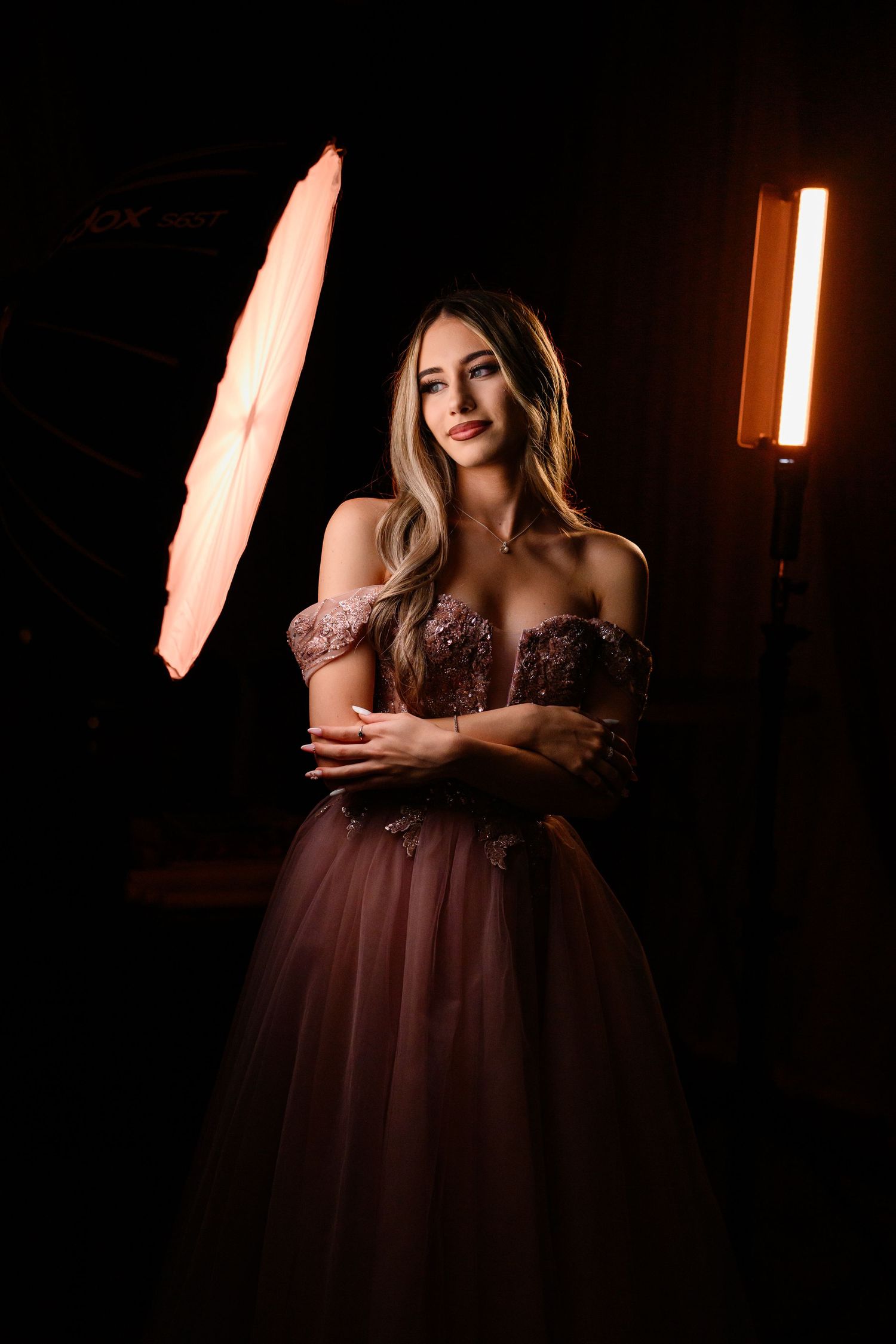 A woman in a dusty rose off-shoulder ball gown poses elegantly in a dark photography studio with professional lighting.