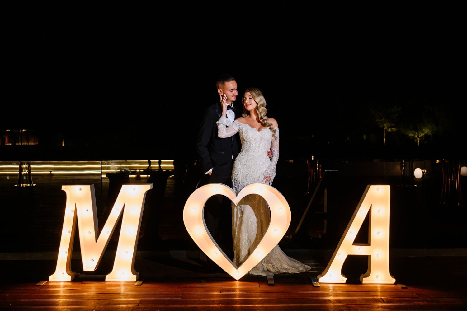 Newlyweds pose between illuminated M and A letters with glowing heart at outdoor night wedding reception.