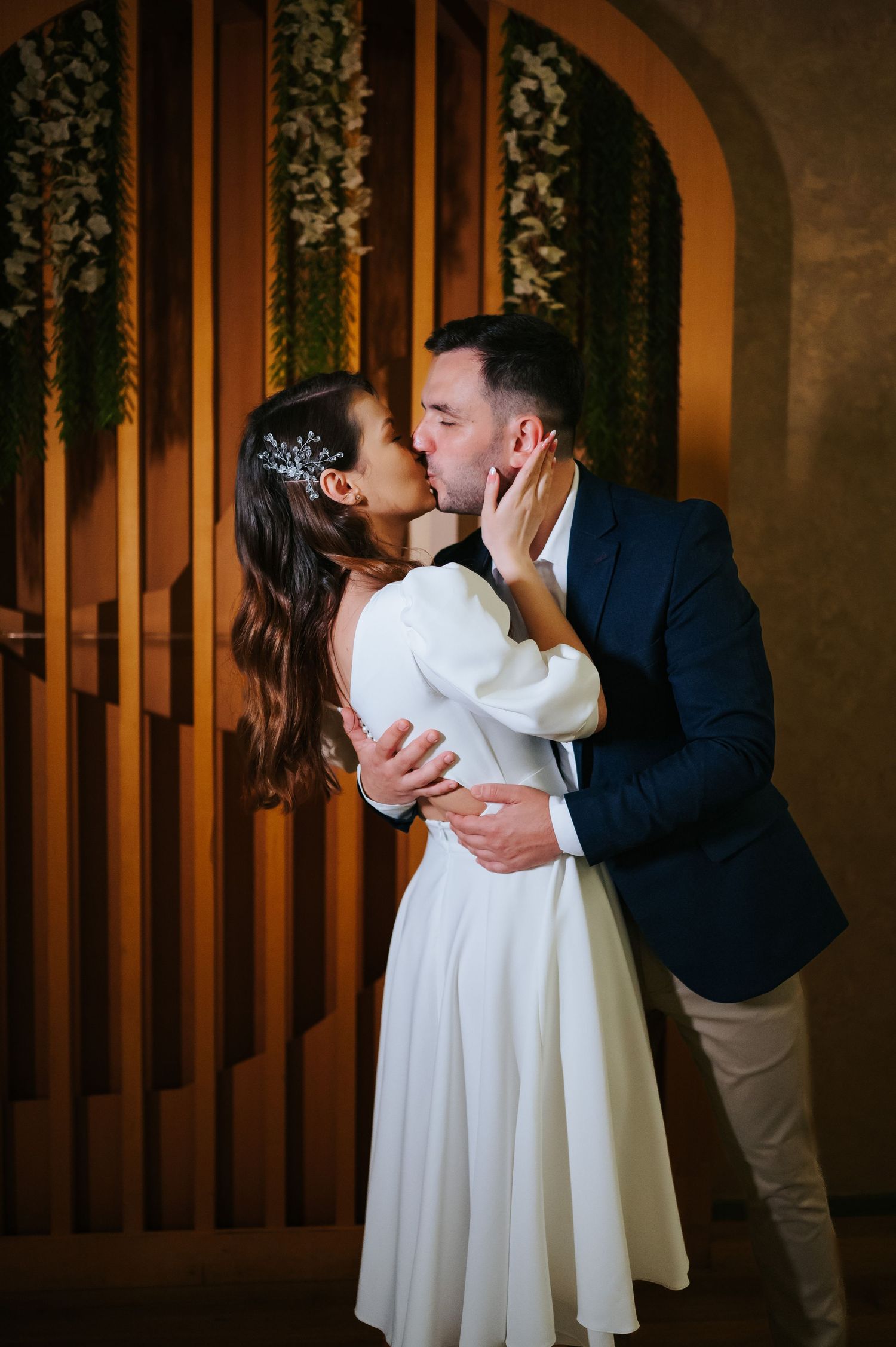 A bride and groom share a romantic kiss, she in a white dress with hair accessory, he in a navy suit.