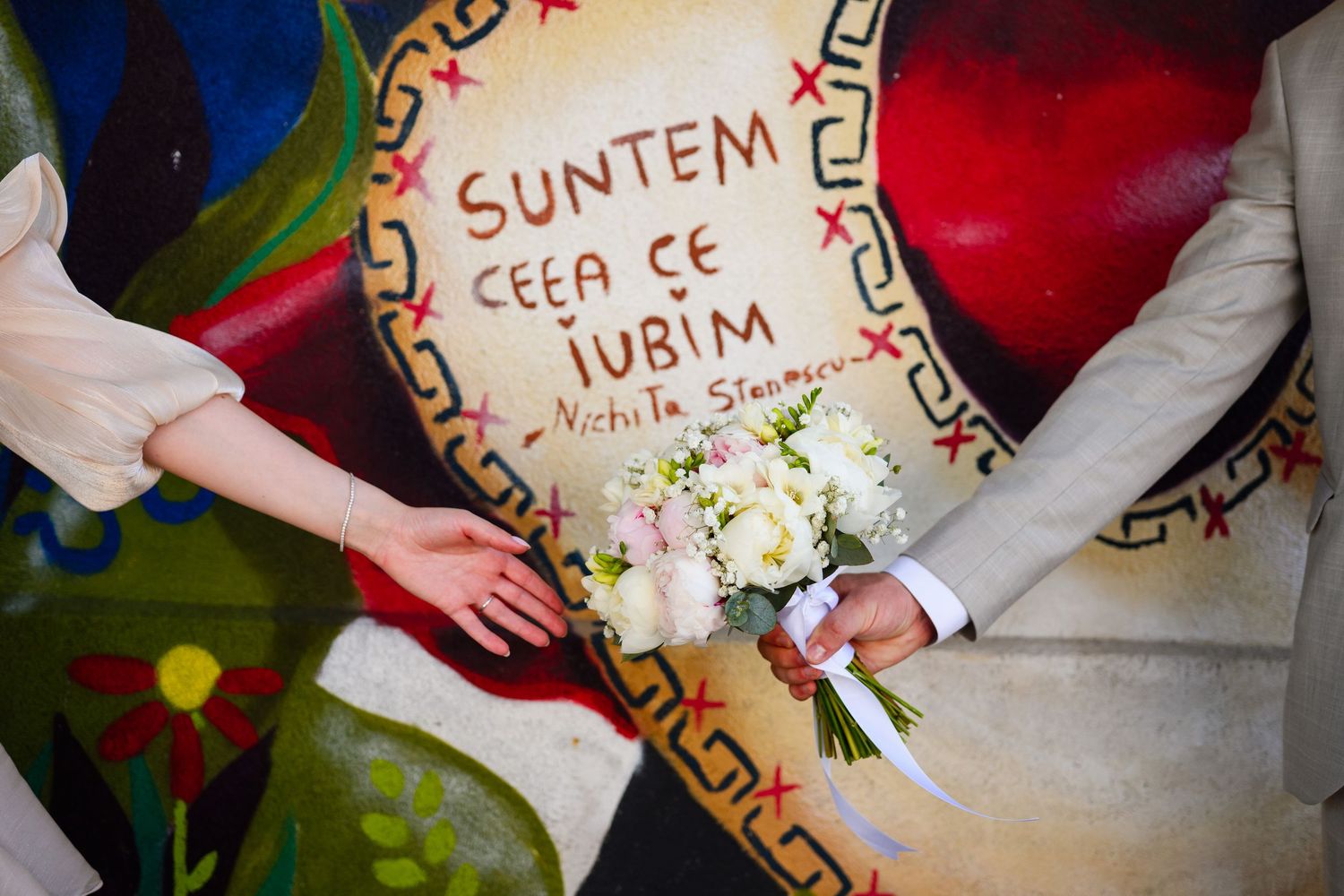 Bride and groom holding a white bouquet in front of a colorful mural with Romanian quote by Nichita Stanescu.