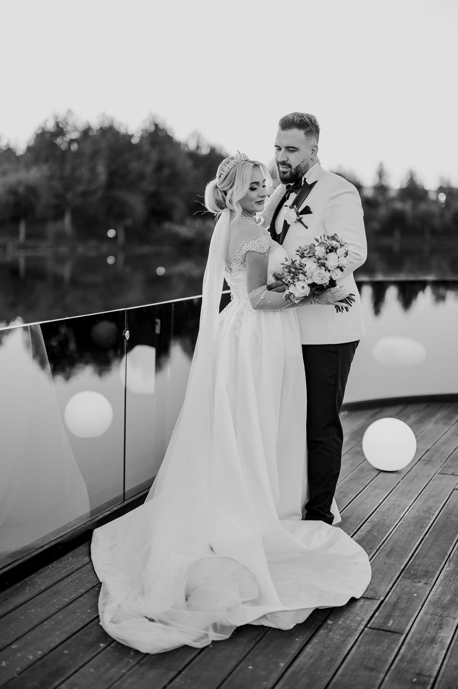 Black and white wedding photo of bride and groom embracing on a dock by a lake, bride holding a bouquet.