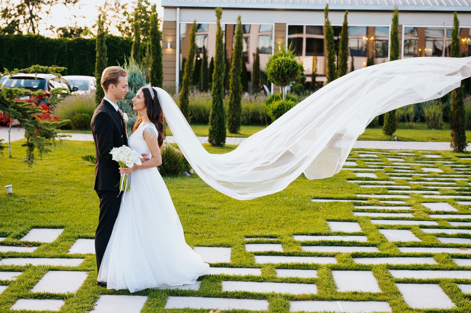 Bride and groom share a romantic moment outdoors, with her flowing veil billowing in the wind at a garden wedding venue.