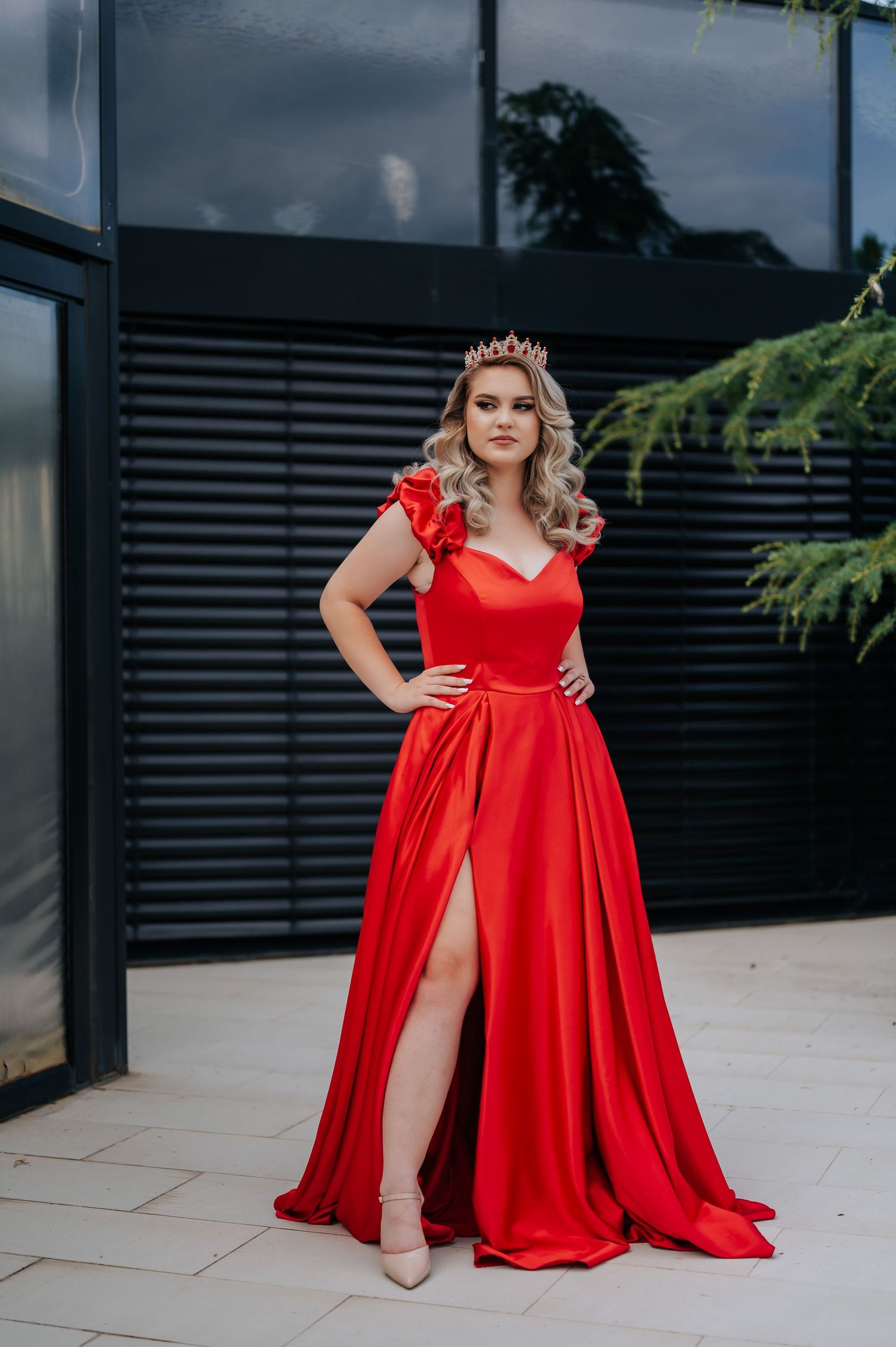 Woman in elegant red satin gown with ruffle shoulders and thigh slit, wearing a crown, posing outdoors.