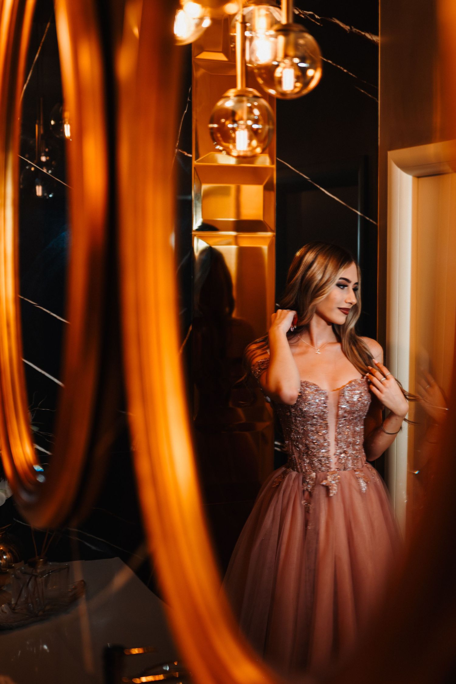 Elegant woman in sparkly pink ballgown poses near ornate gold mirror under warm ambient lighting.