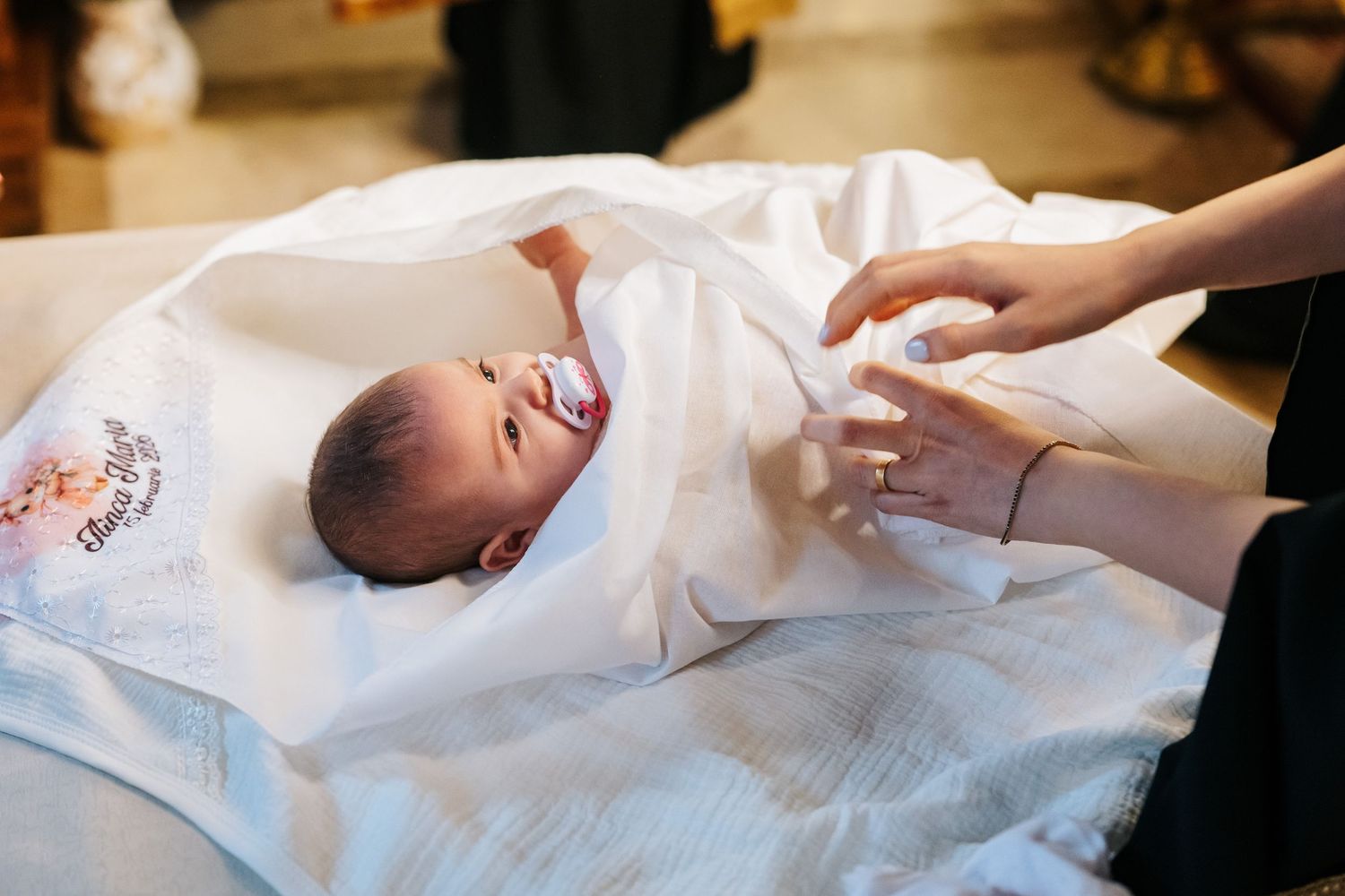 Newborn baby with pacifier resting on white ceremonial baptism cloth held by adult hands.