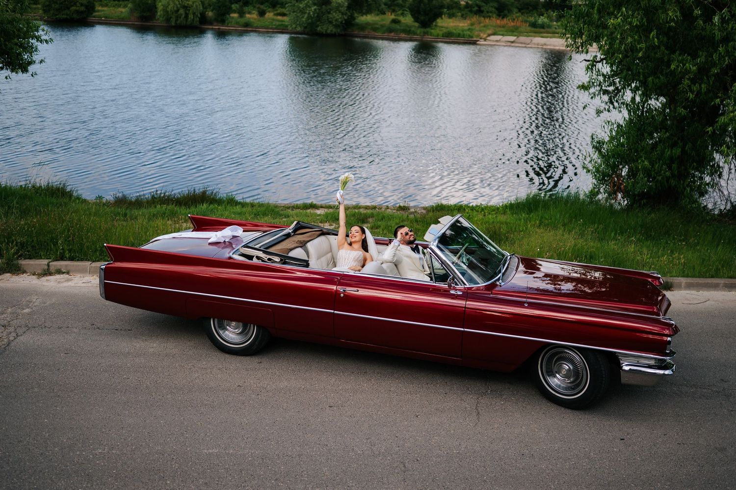 Bridal party rides in stunning red vintage Cadillac convertible along scenic lakeside road on wedding day.