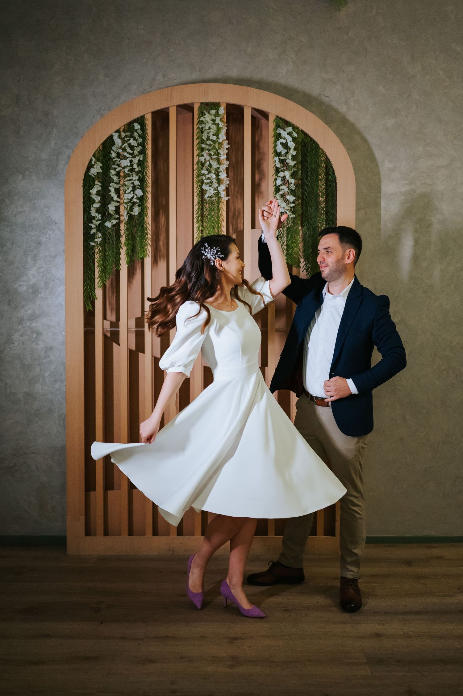 Bride in white midi dress twirls with groom in navy blazer dancing before an arched wooden backdrop.