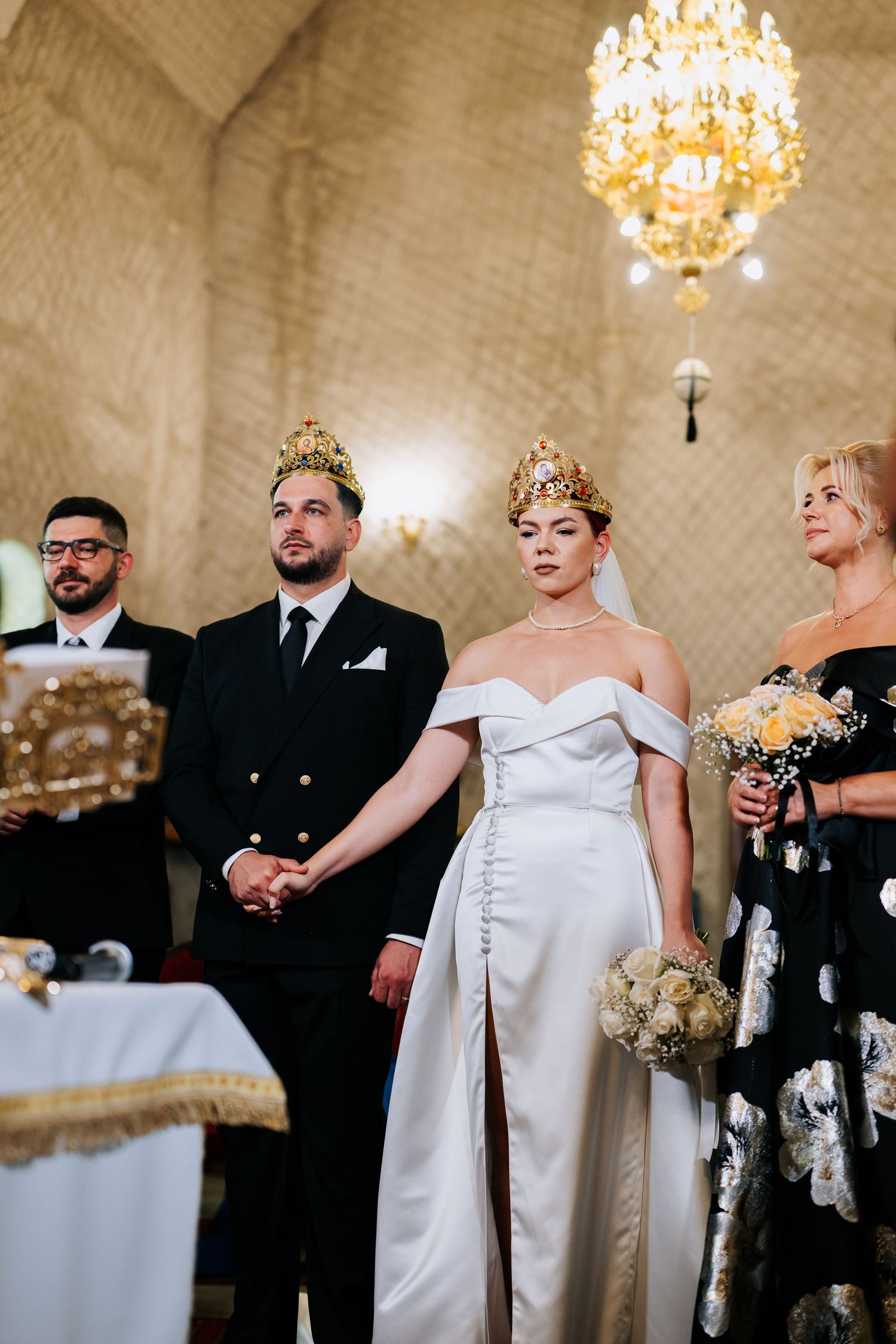 Bride and groom wearing gold crowns hold hands during an Orthodox wedding ceremony in an elegant church.