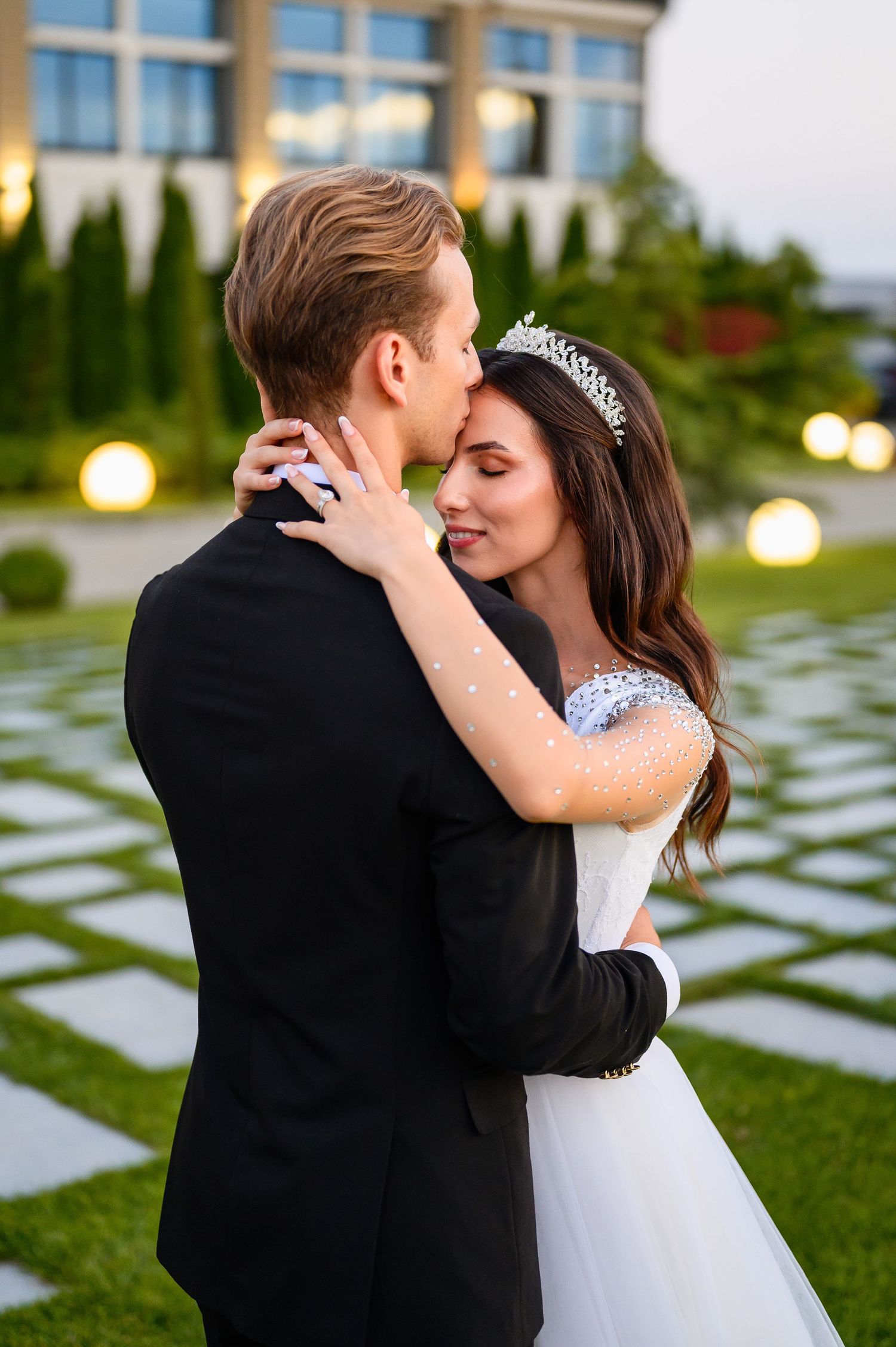 Groom kissing bride wearing tiara as they embrace in softly lit garden at dusk.
