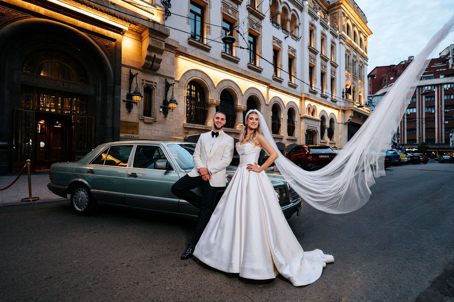 Elegant bride and groom pose beside a vintage teal car on a city street at dusk, veil flowing.