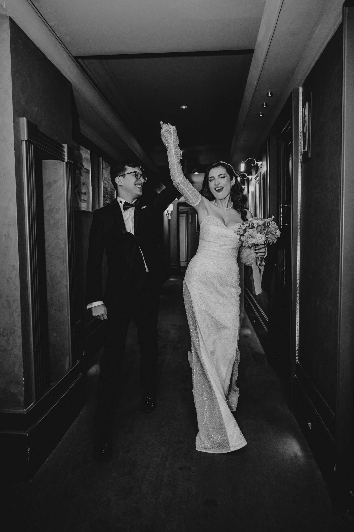 Black and white photo of joyful bride and groom walking through elegant hotel corridor.
