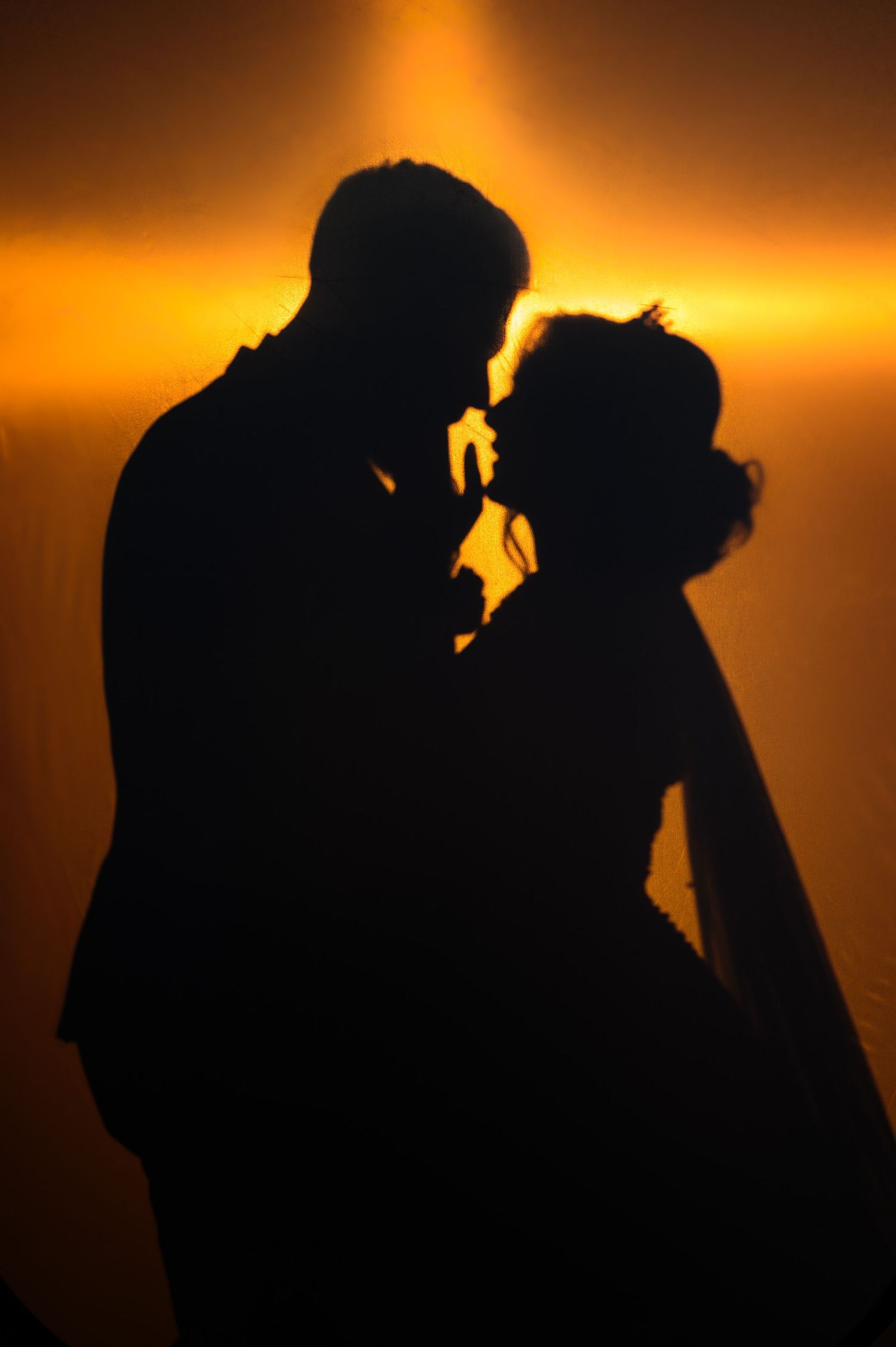 Romantic silhouette of a couple about to kiss against a warm golden orange sunset backdrop.