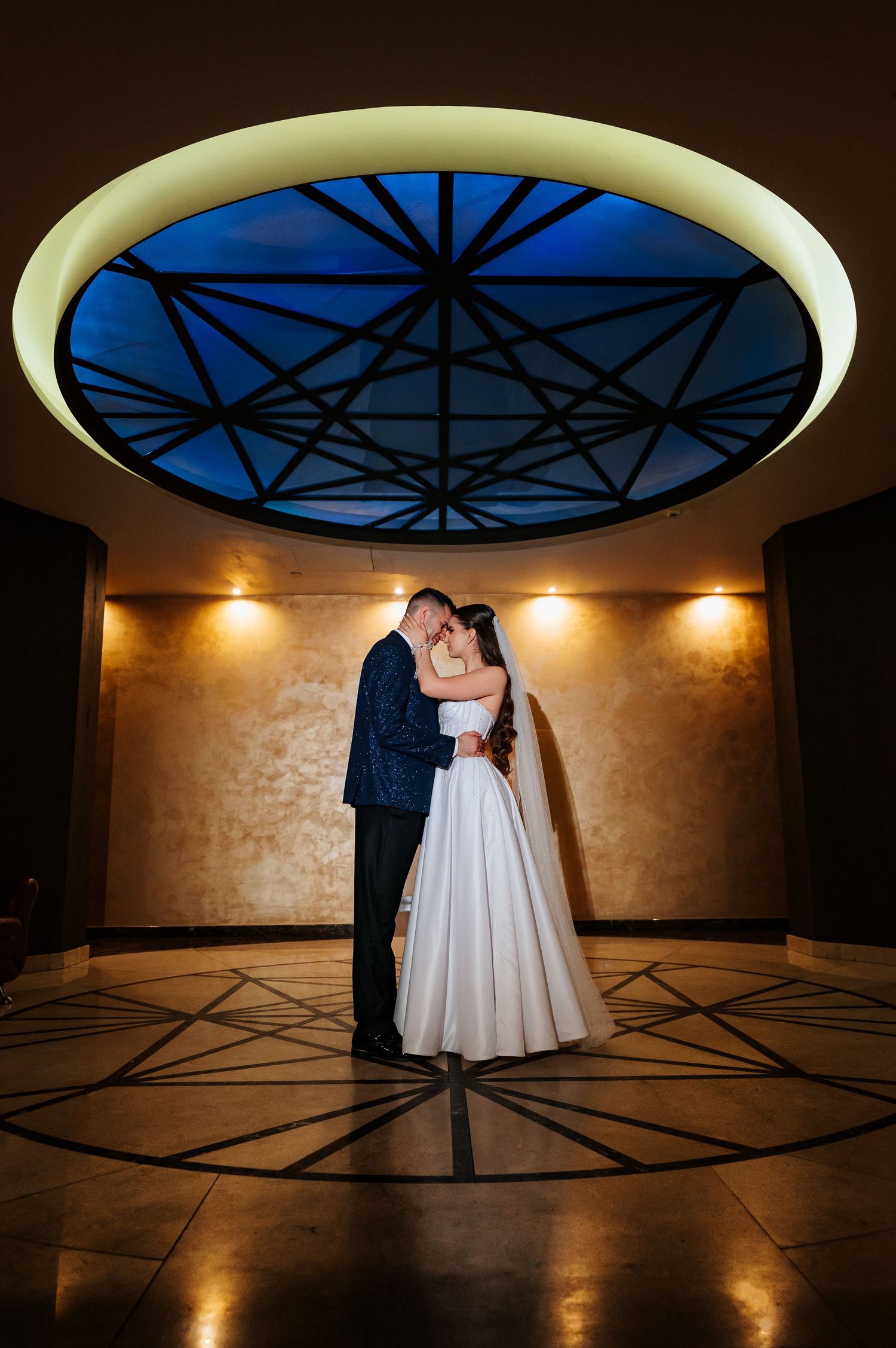 Bride and groom embrace under glowing circular skylight in elegant wedding venue.