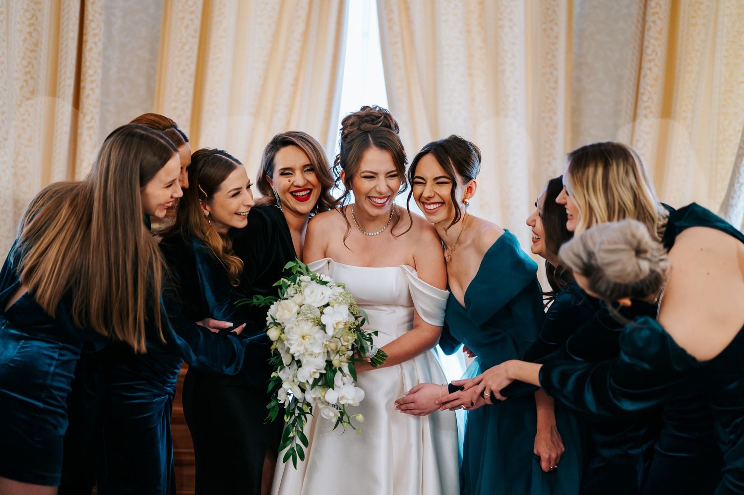 Smiling bride holding white bouquet surrounded by bridesmaids in teal dresses at wedding.