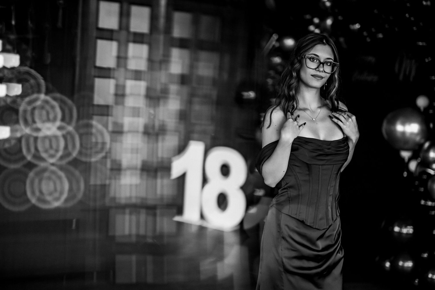 Black and white photo of a woman in an elegant strapless dress posing near a large number 18 sign.