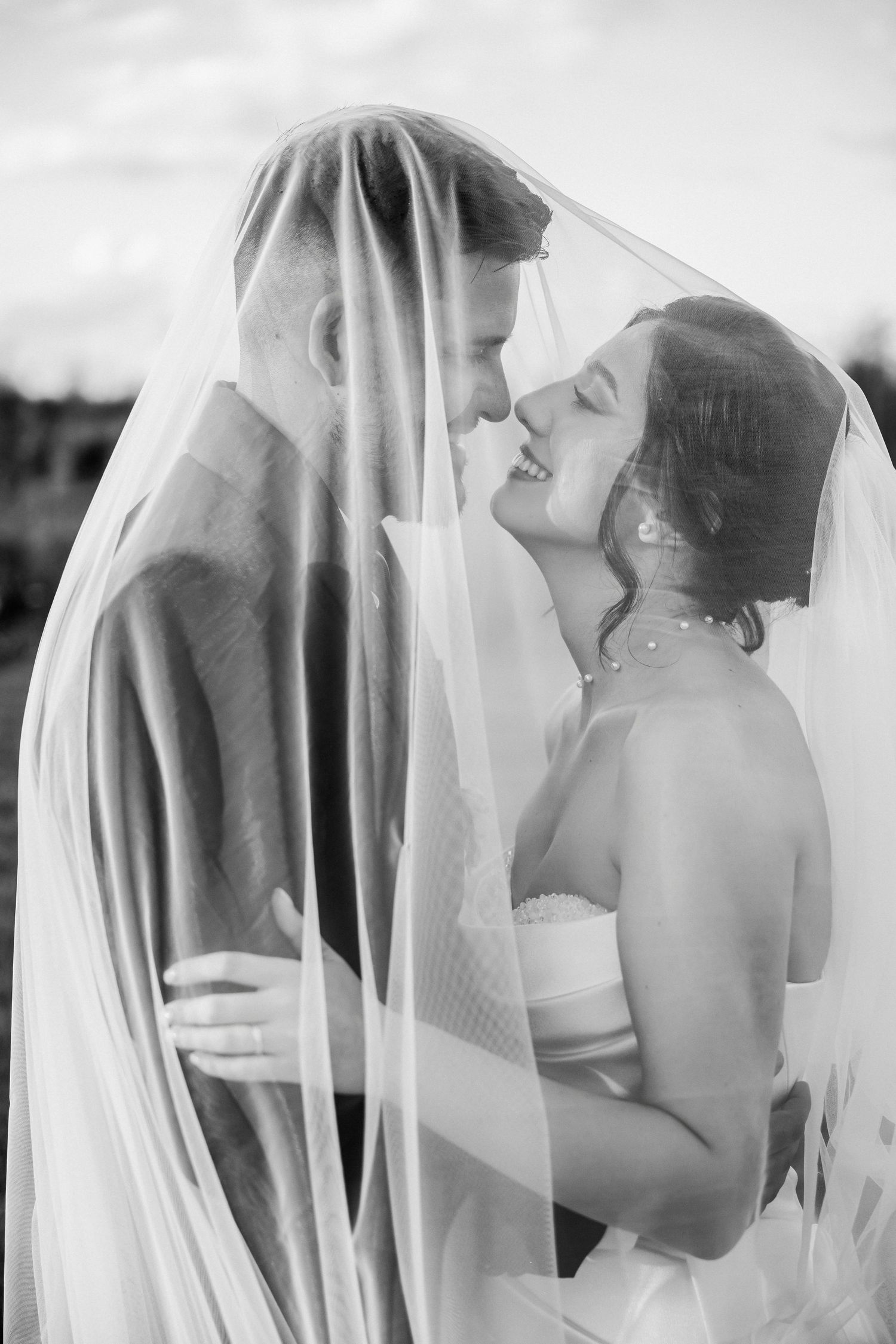Black and white photo of bride and groom sharing a romantic moment under a flowing wedding veil.