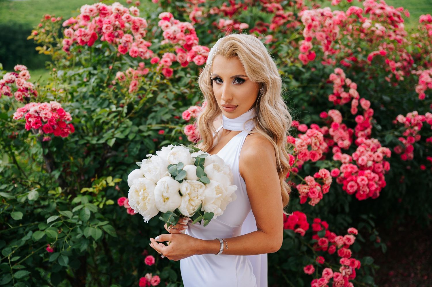 Blonde bride in white dress holding white rose bouquet poses before blooming pink rose bushes outdoors.